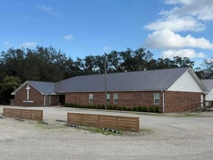 A single-story brick church building with a gray metal roof, a white cross on the front wall, and a wooden parking fence.