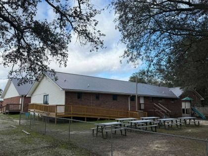 A one-story brick building with a metal roof, featuring a wooden wheelchair ramp, set in a yard with picnic tables.