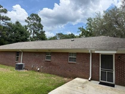 A red brick exterior wall of a one-story house with a grey shingled roof, a back door with a screen, and a concrete patio.