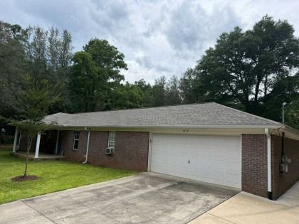 A single-story, brick-faced ranch-style house with a light gray shingled roof, white garage door, and a grassy front lawn.