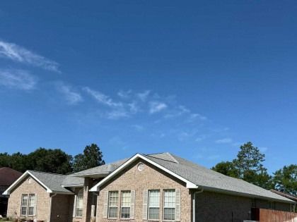 A tan brick, single-story house with a gray shingle roof under a clear blue sky.