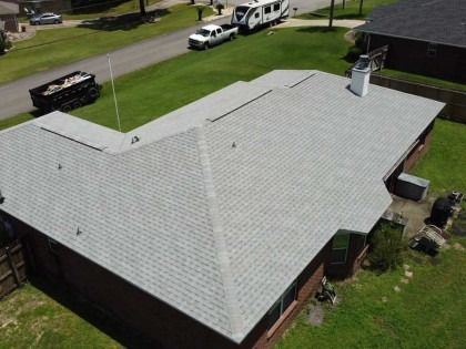 An aerial view of a gray-shingled roof on a brick house, with a trailer parked on the street in front of the yard.
