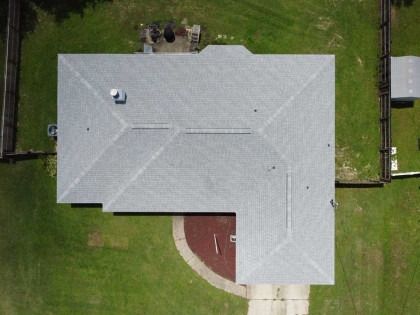 An aerial view of a gray-shingled L-shaped roof on a house, surrounded by a grassy lawn with a fence and a walkway.