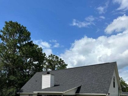 A white house with a gray shingle roof and a central chimney set against a blue sky with clouds and tall pine trees.