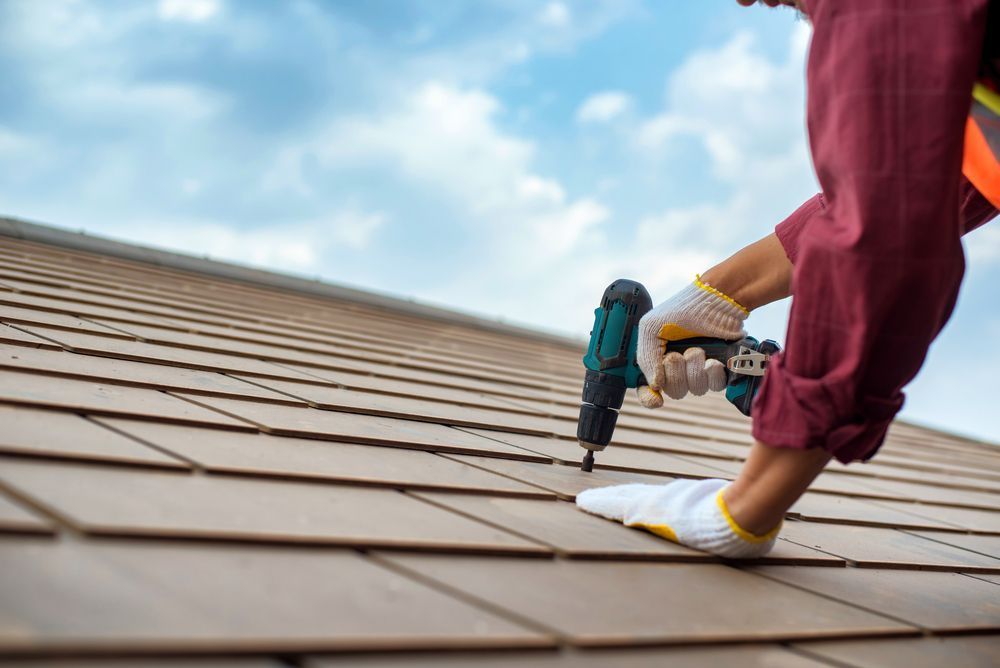 A worker in a maroon long-sleeved shirt and white work gloves uses a power drill to install brown roof tiles.