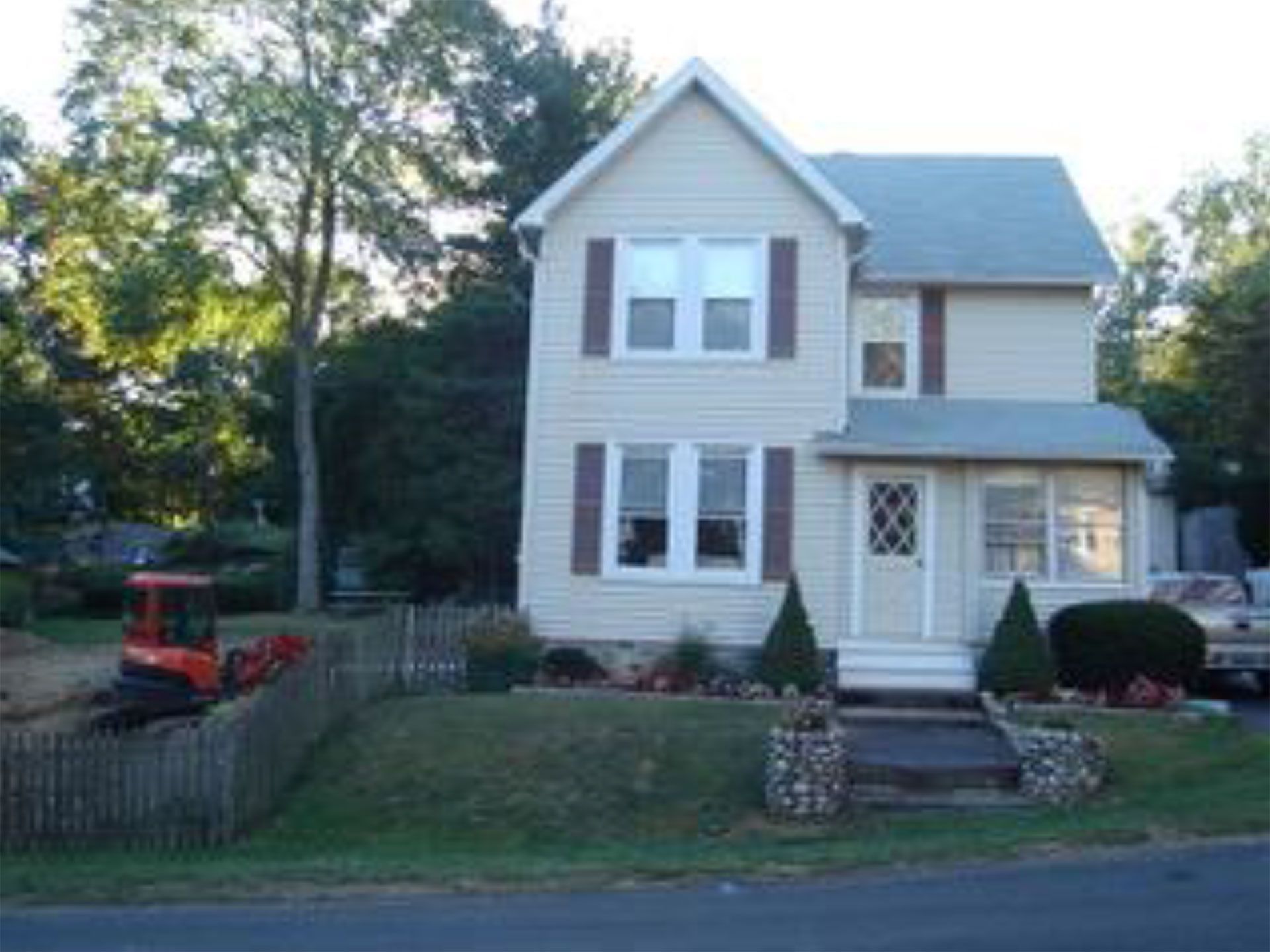 Two-story beige house with brown shutters and a small stone porch. A small excavator and fence are in the front yard.