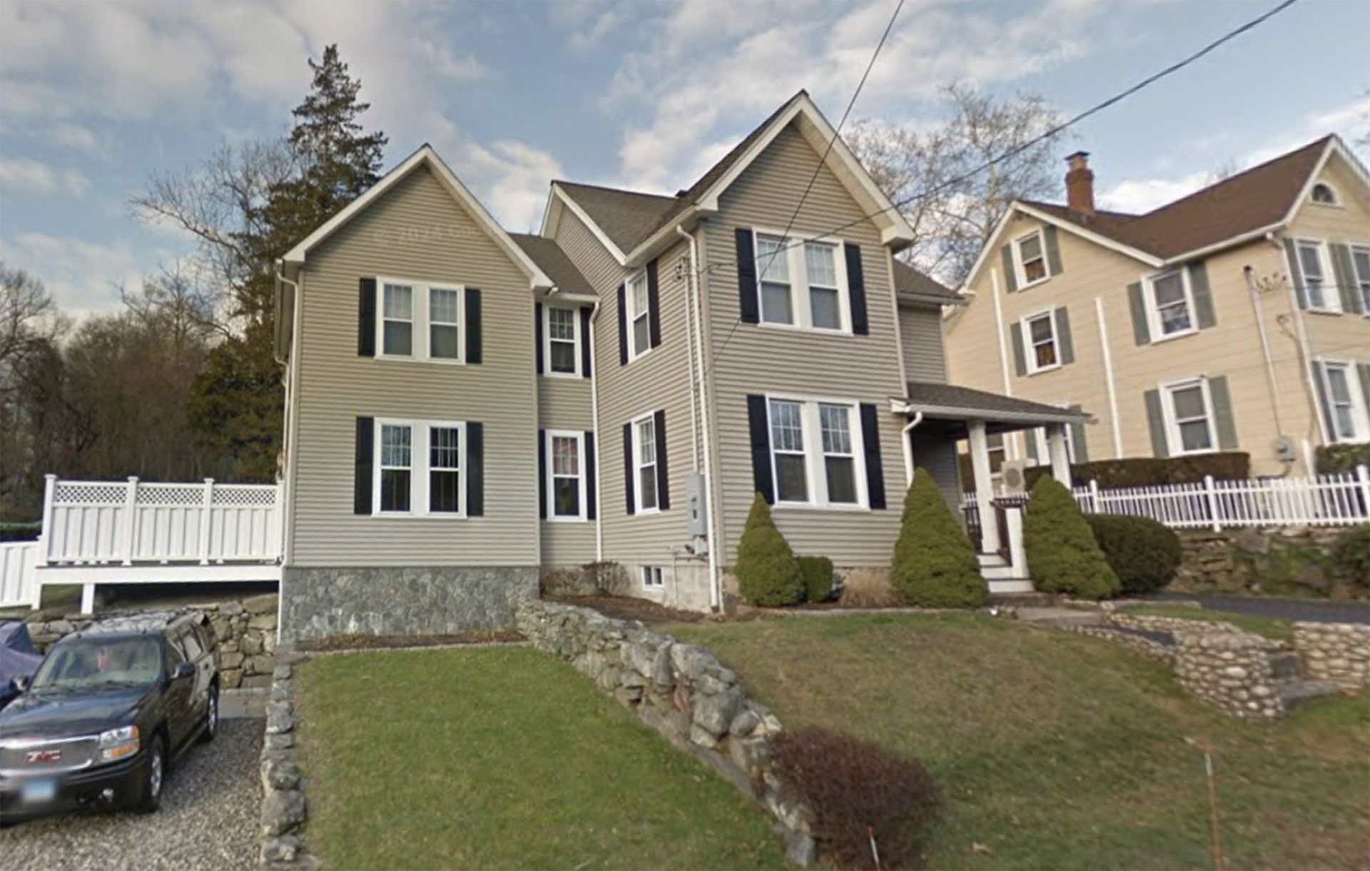 Two-story beige house with black shutters, on a sloped lawn. Stone retaining wall and car in the front.
