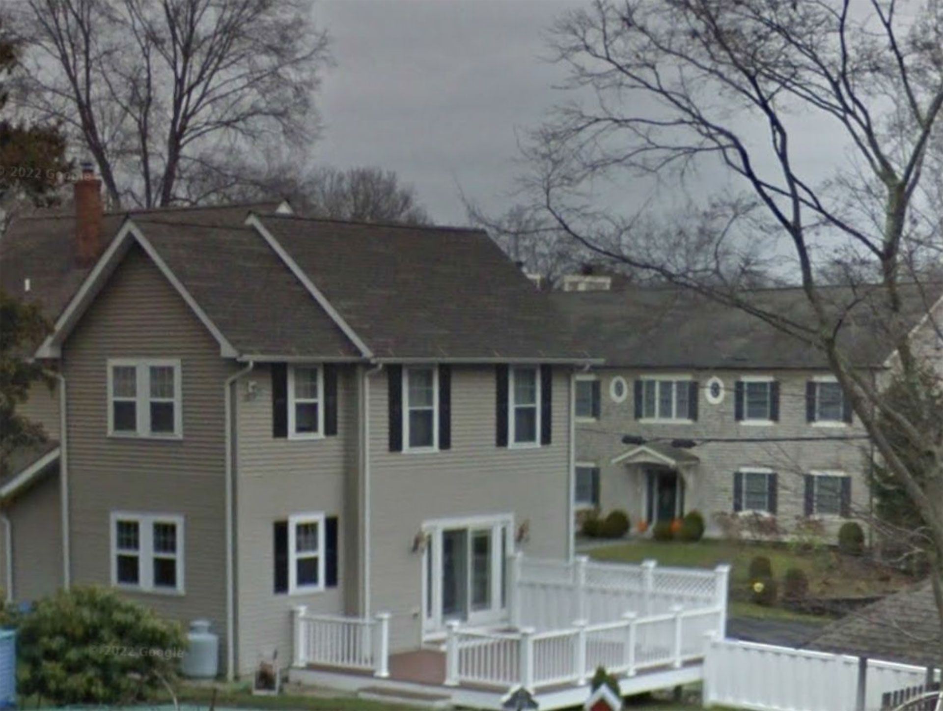 Two-story house with gray roof and siding, white deck, and black shutters, trees in background.