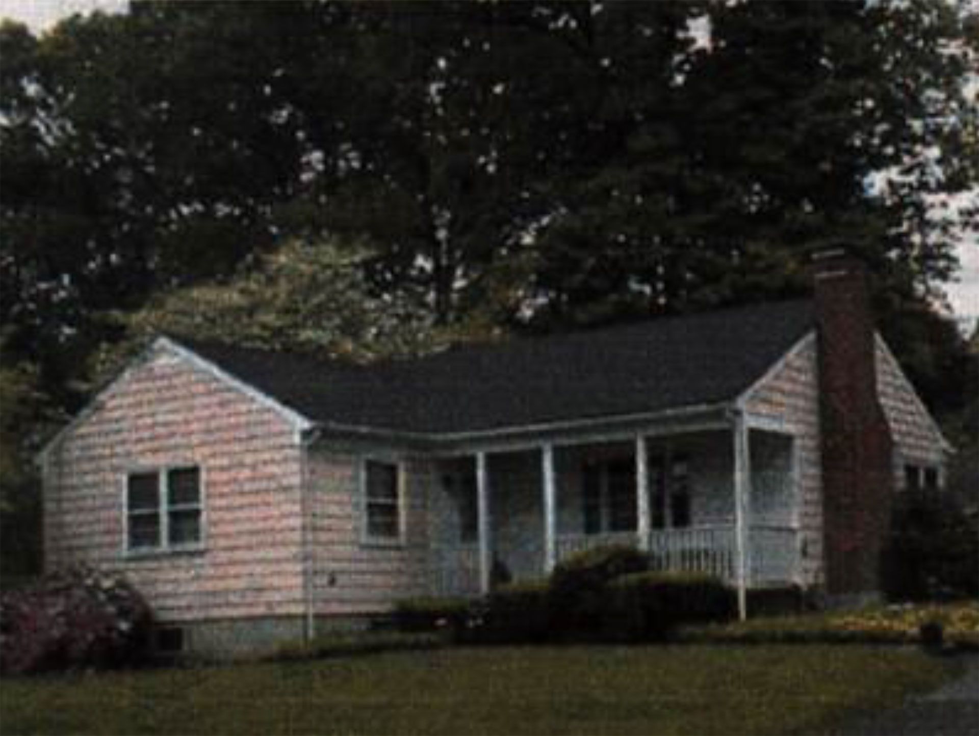 A small, pink-sided bungalow with a porch and brick chimney, set against a backdrop of trees.
