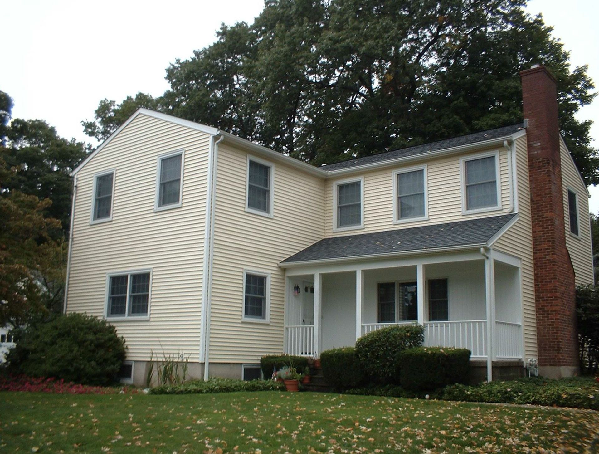 Two-story house with light siding, dark roof, brick chimney, and porch, set in front of green lawn and trees.