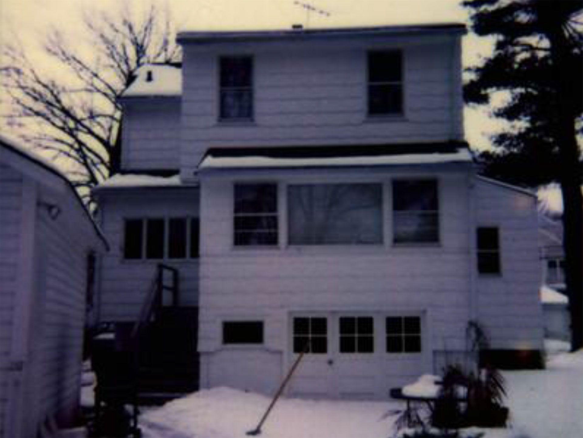 White two-story house in winter with snow-covered yard. Broom and some plants are visible outside.