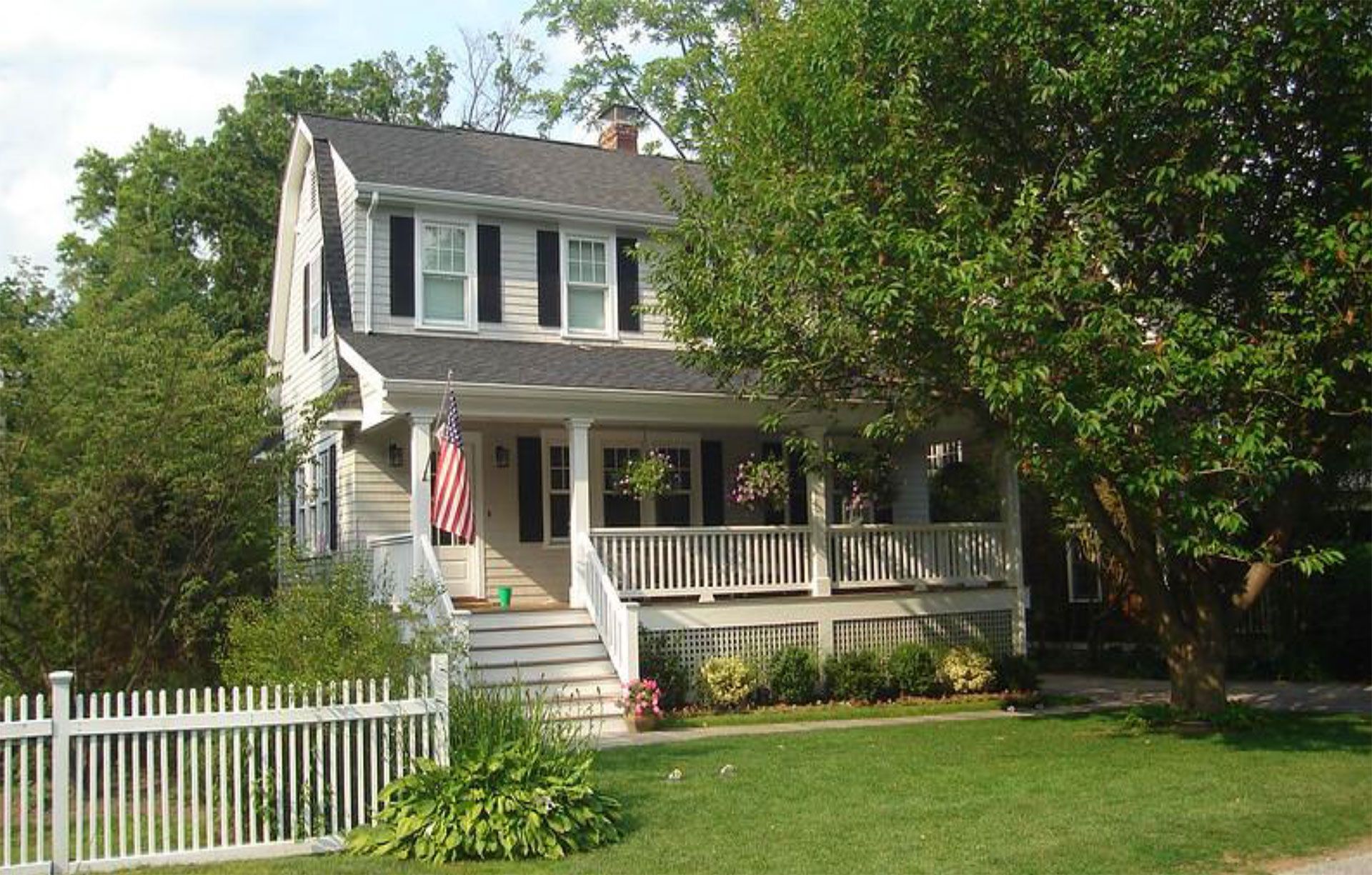 Two-story house with a white picket fence, covered porch, and American flag; green lawn and trees.