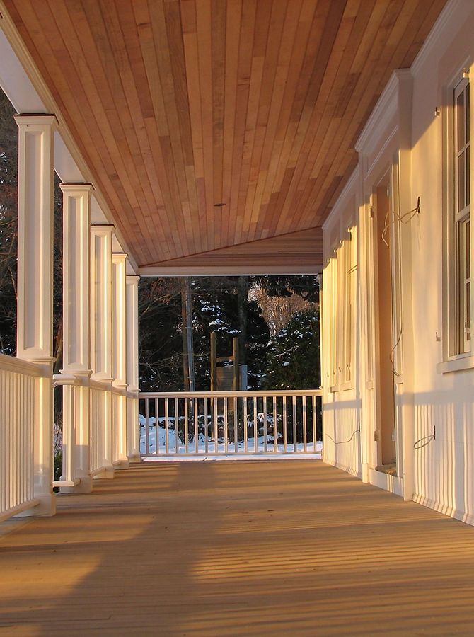 Wooden porch with white columns, railing, and ceiling. Snow outside, illuminated by sunlight.