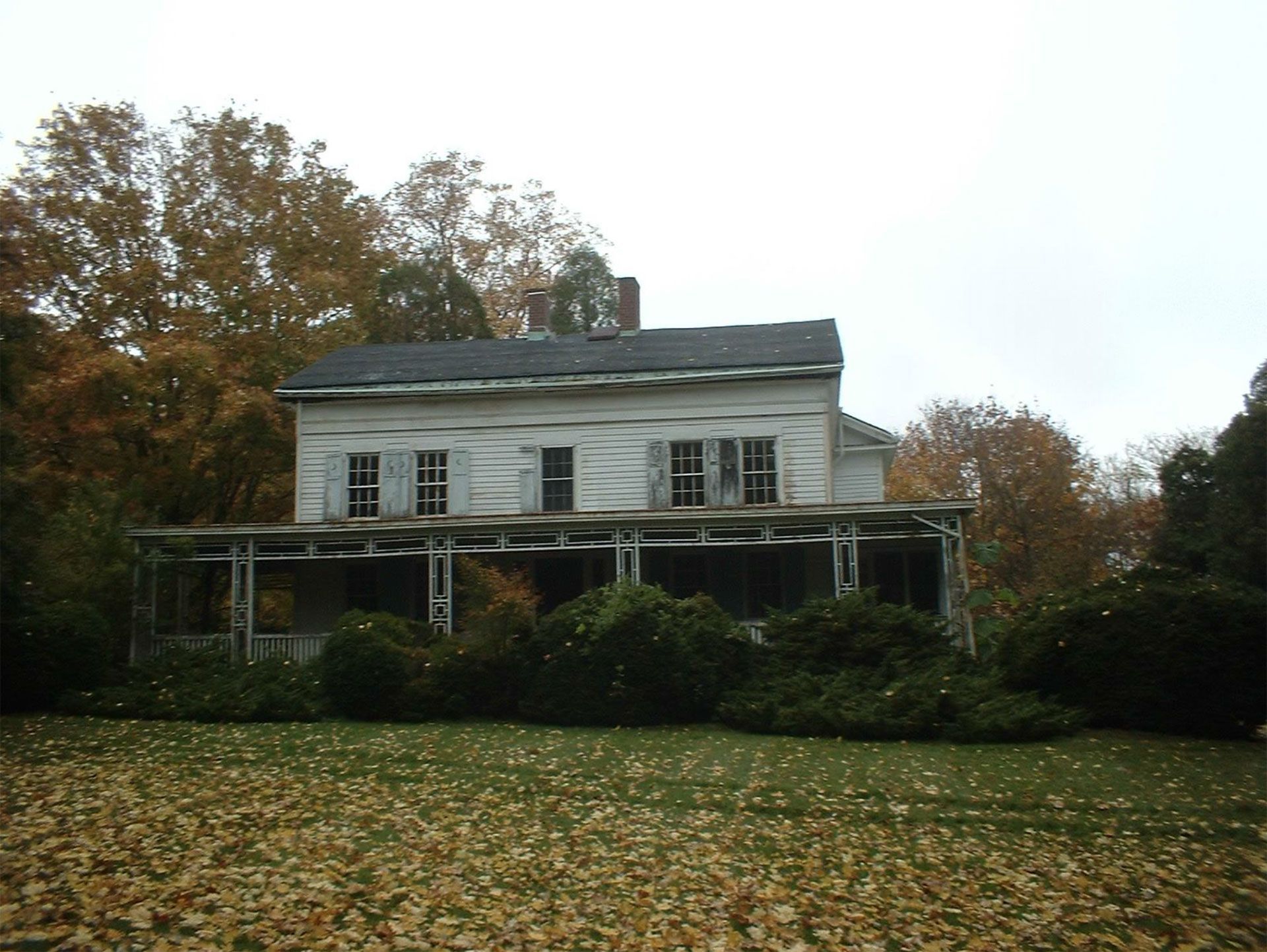 Dilapidated white two-story house with shutters, overgrown porch, surrounded by fall foliage.