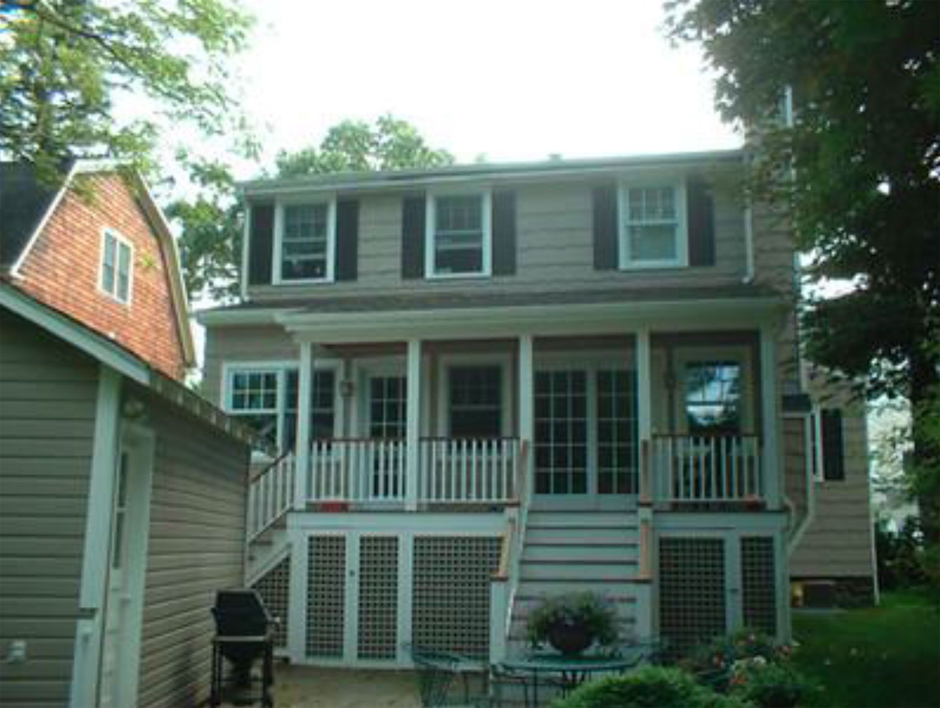 Tan two-story house with porch and black shutters, shed on the left, trees surround.