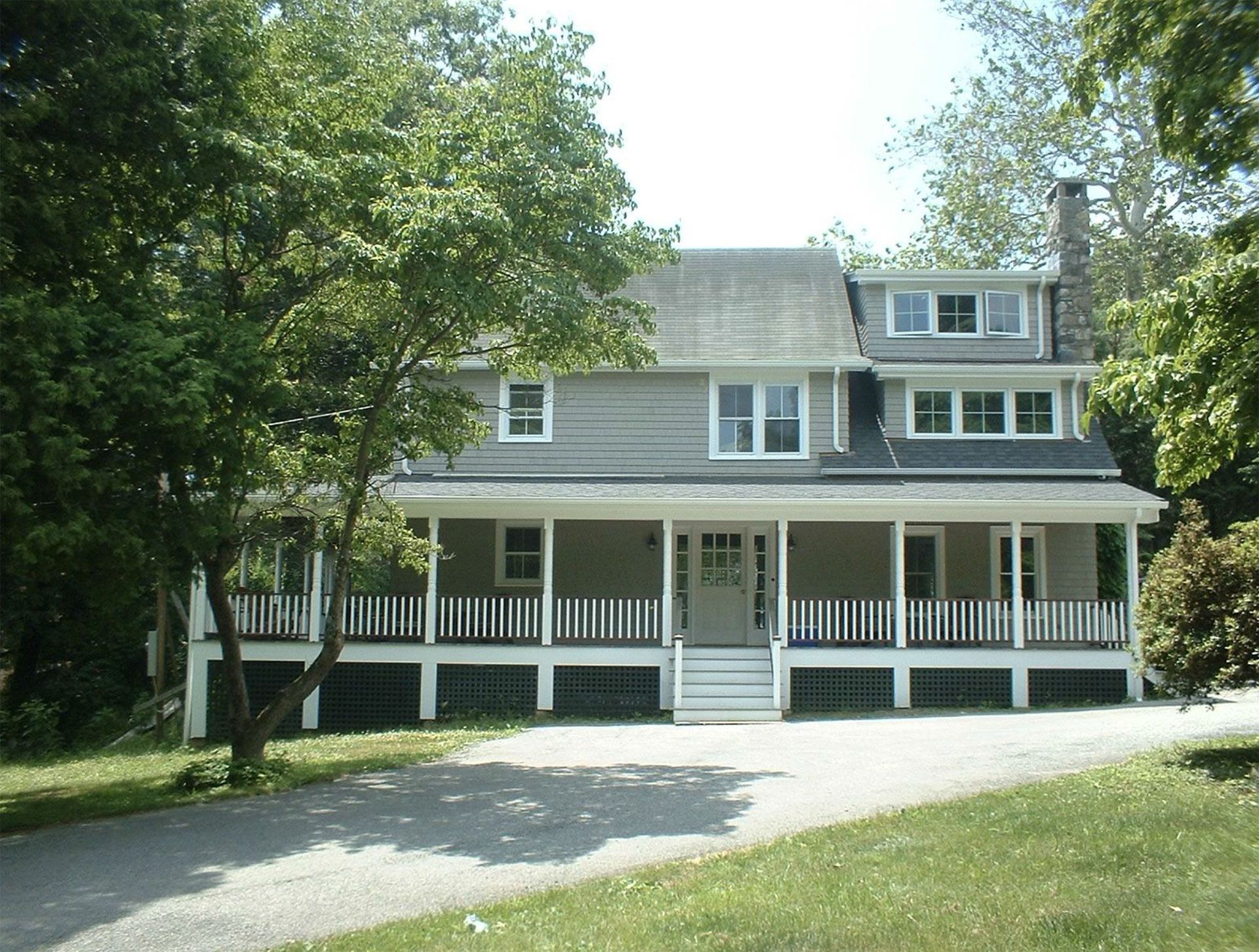 Two-story light gray house with wraparound porch and dark railings, set in a tree-lined driveway.