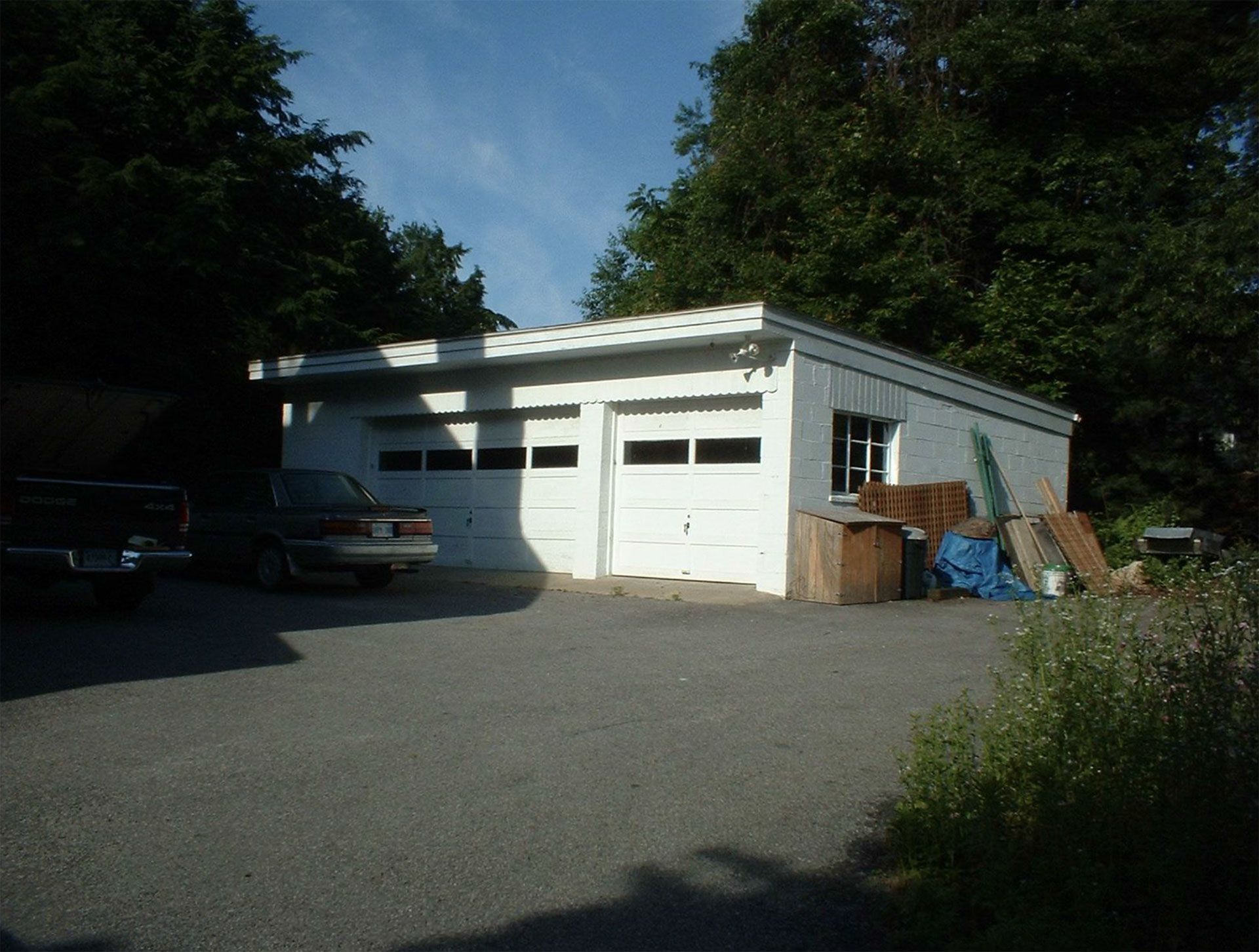 White two-car garage with overhead doors and parked vehicles in a gravel driveway, surrounded by trees.