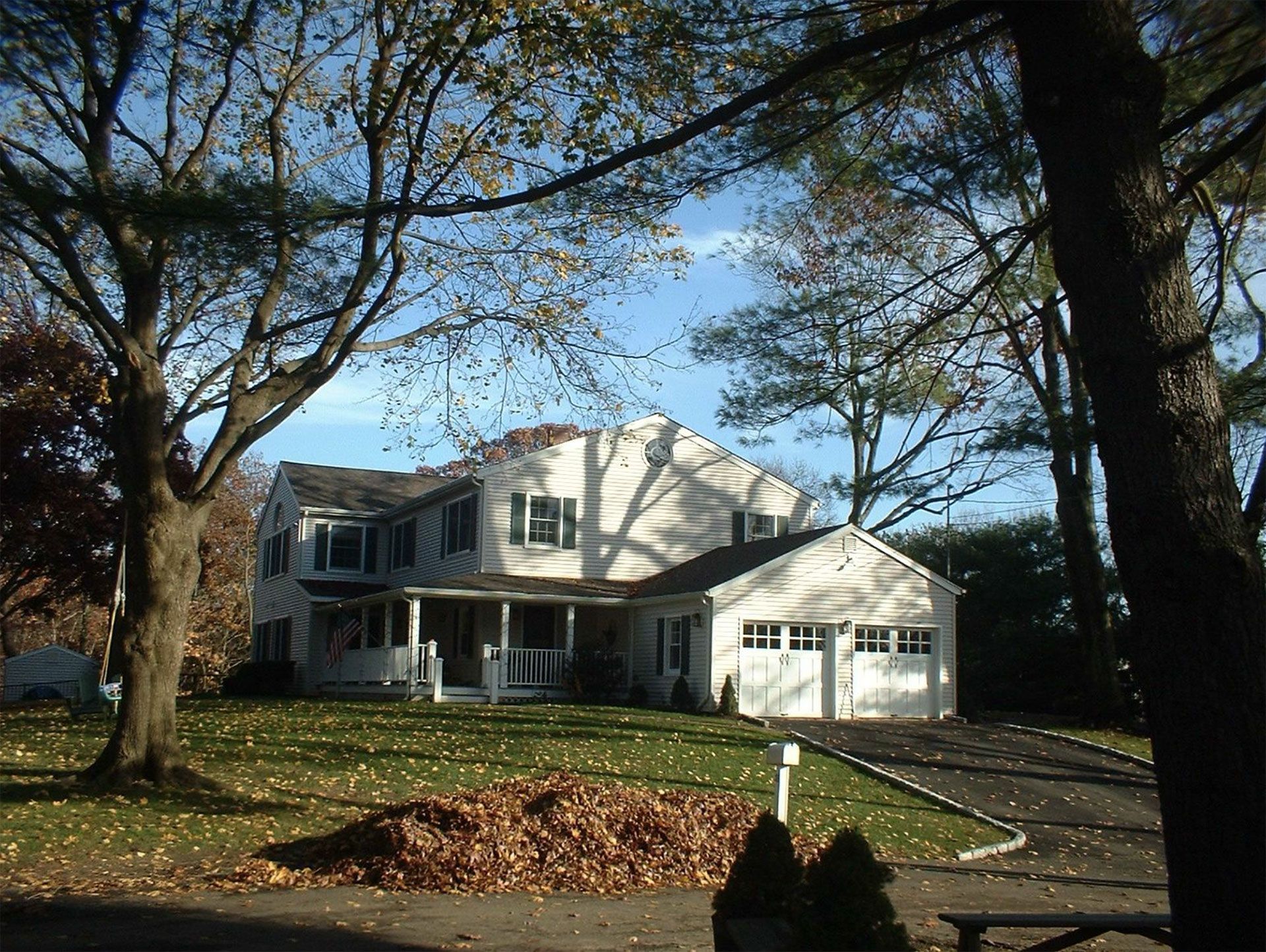 Two-story white house with attached garage, trees, and a pile of leaves on a sunny day.