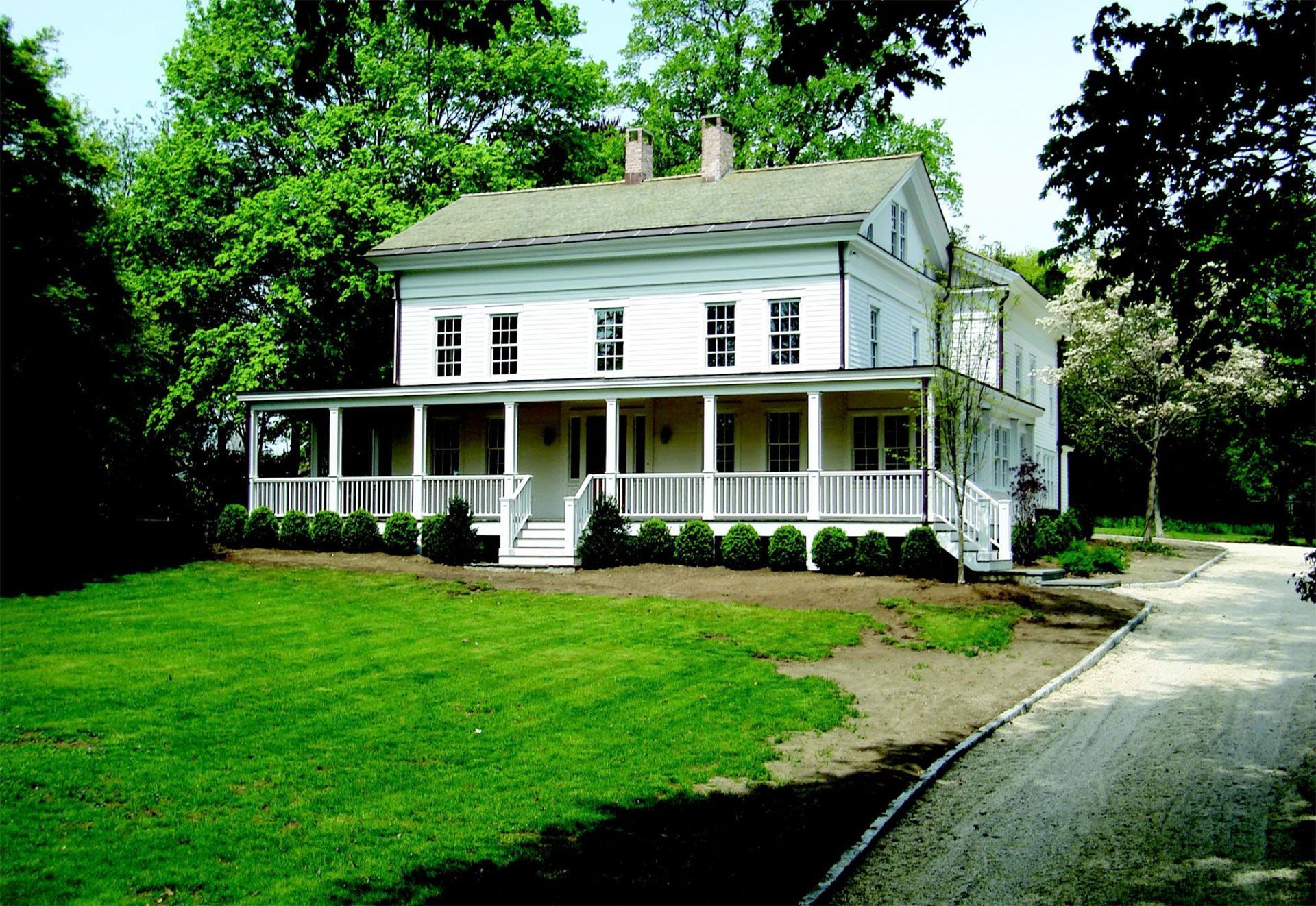 White two-story house with a porch and green lawn, trees surrounding, and a pathway to the right.