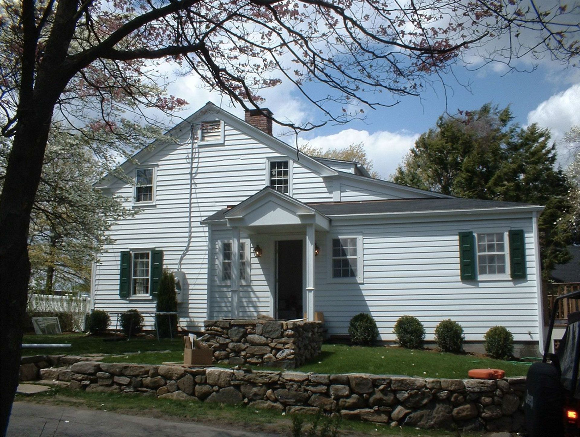 White house with green shutters, stone wall, blue sky.