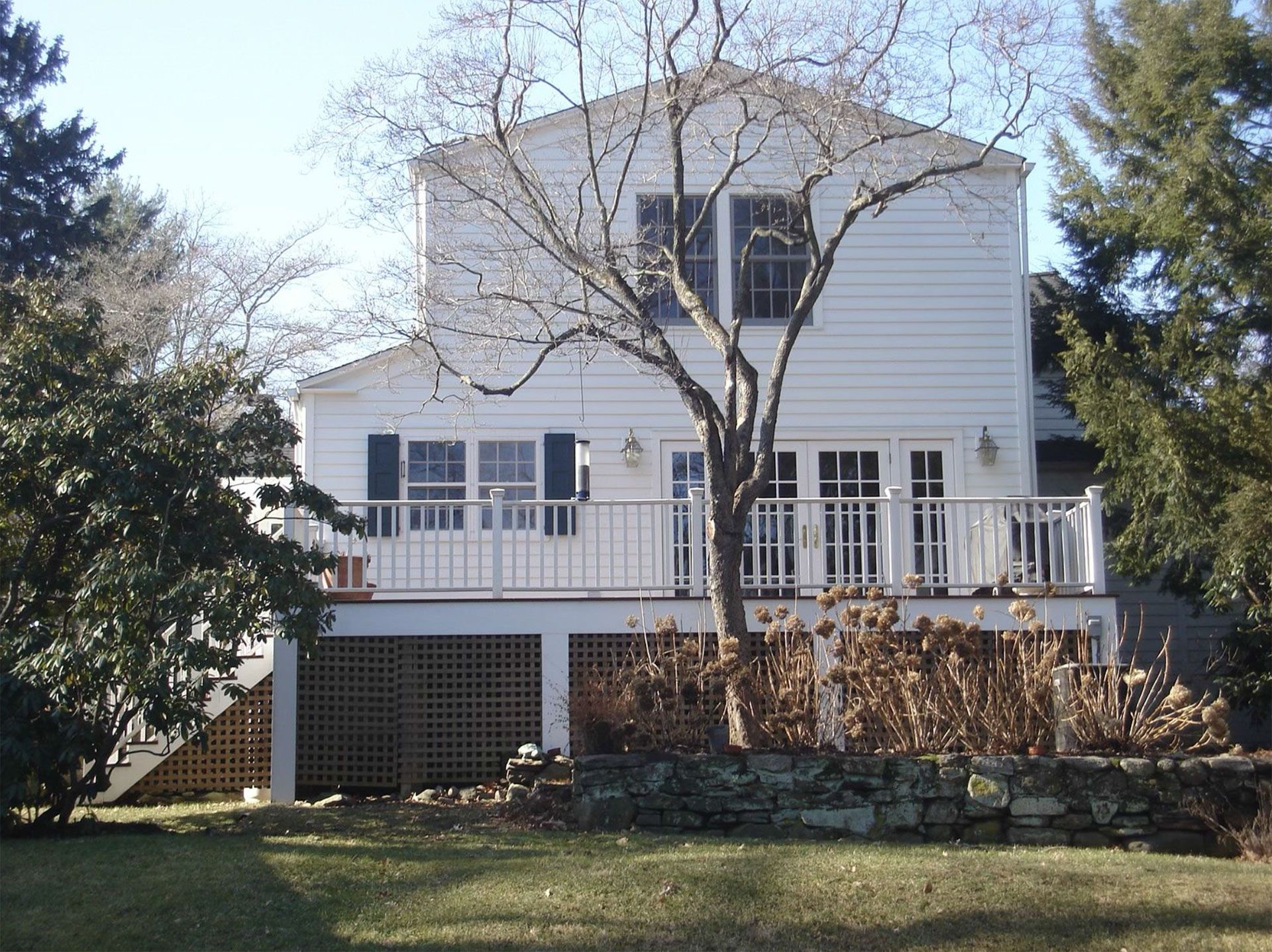 Two-story white house with a deck, small stone wall, and bare trees on a sunny day.