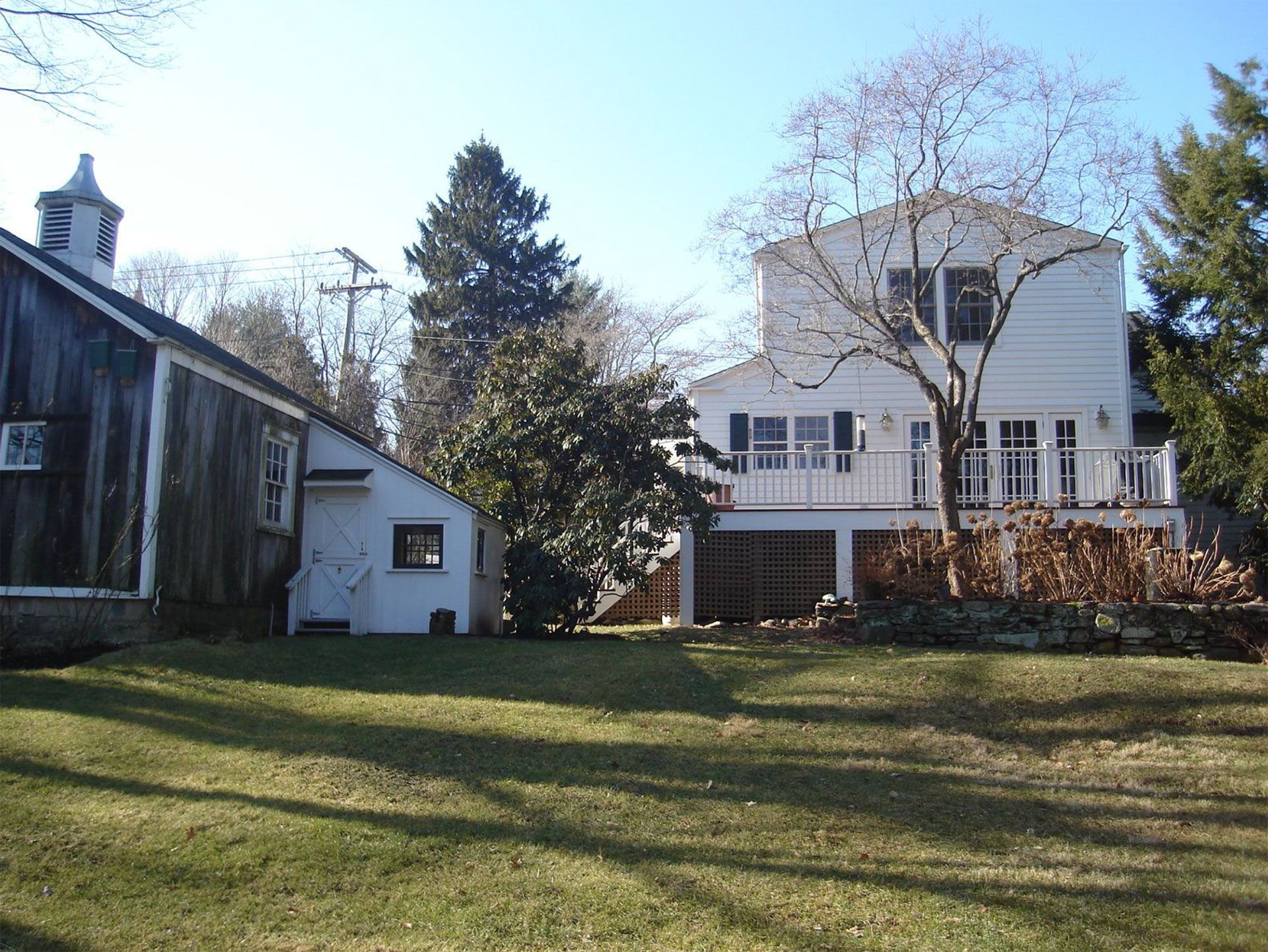 White house with deck and brown barn on a sunny day. Green grass in the foreground.