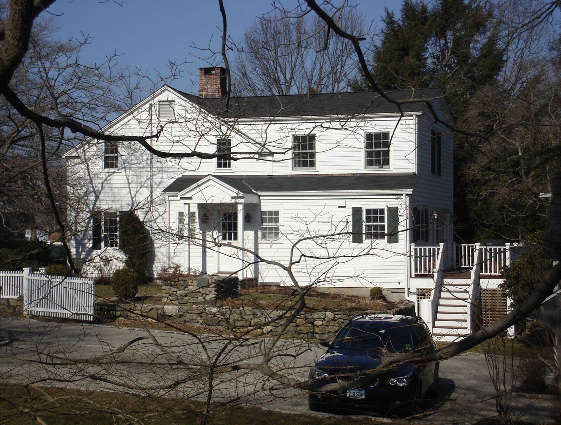 White two-story house with black shutters, a dark SUV in driveway, and leafless tree branches in the foreground.