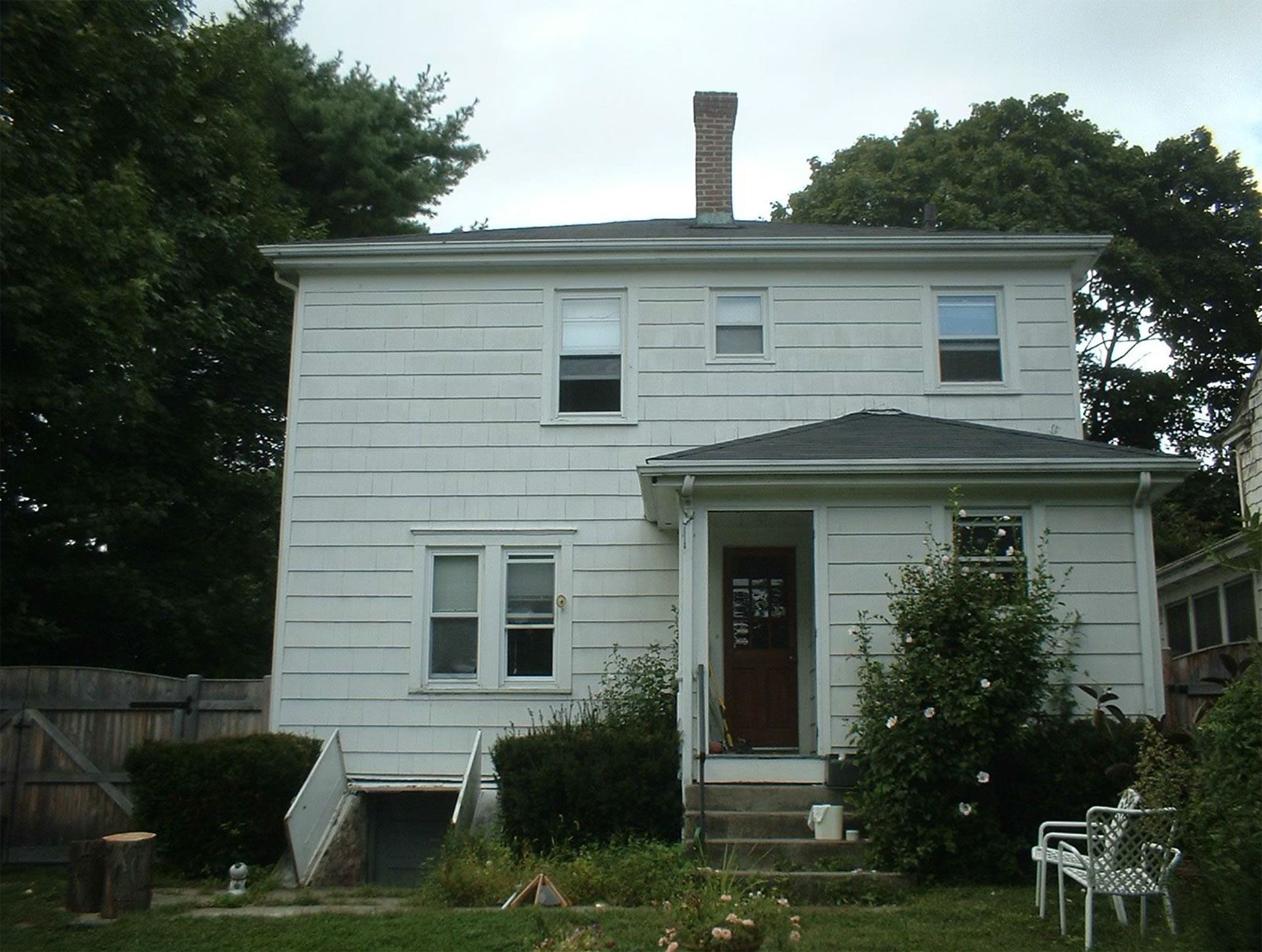 White two-story house with dark roof, chimney, and door. Small porch, grass, and shrubs in front.