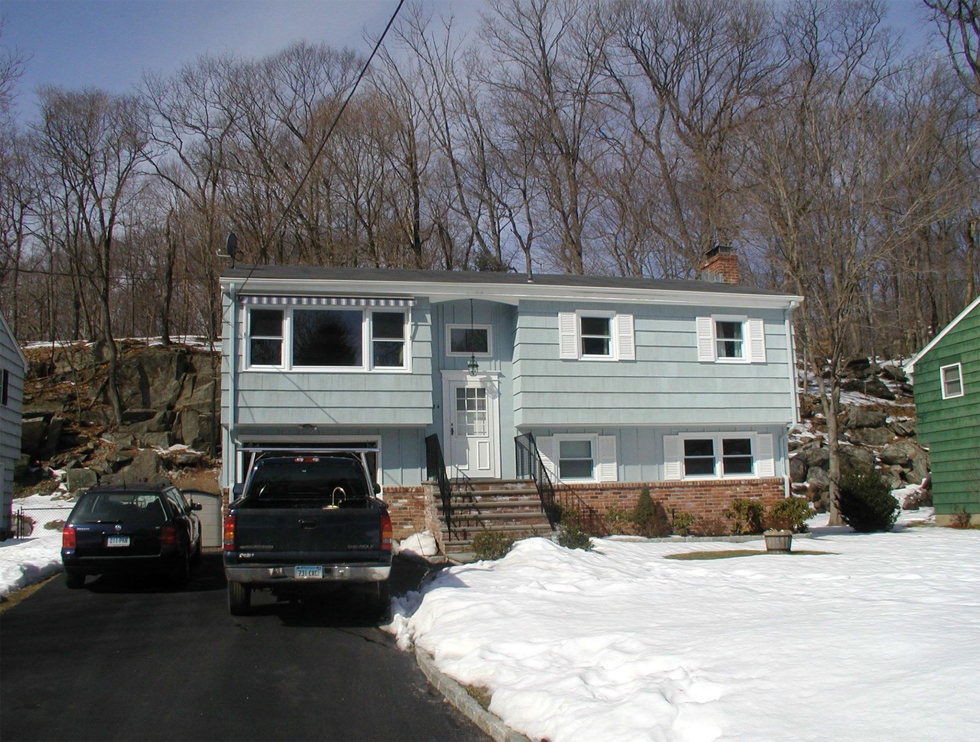 Blue house with open garage, two vehicles parked in front, snow-covered yard, trees in background.