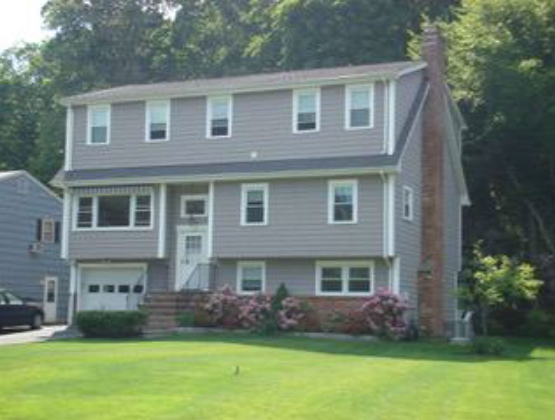 Two-story gray house with garage, chimney, and green lawn. White windows and trim, pink flowers.