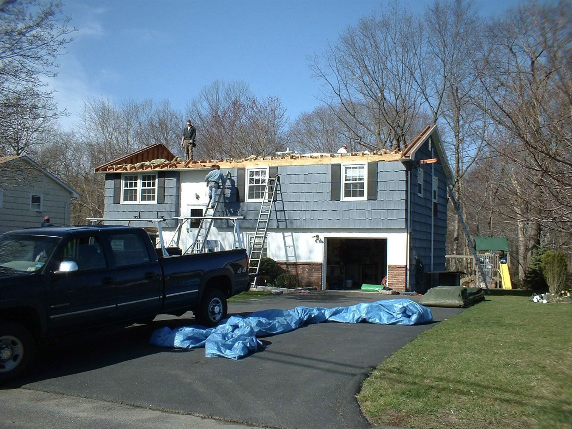Roofers on a blue-sided house replacing the roof; blue tarp on driveway, truck parked in front.