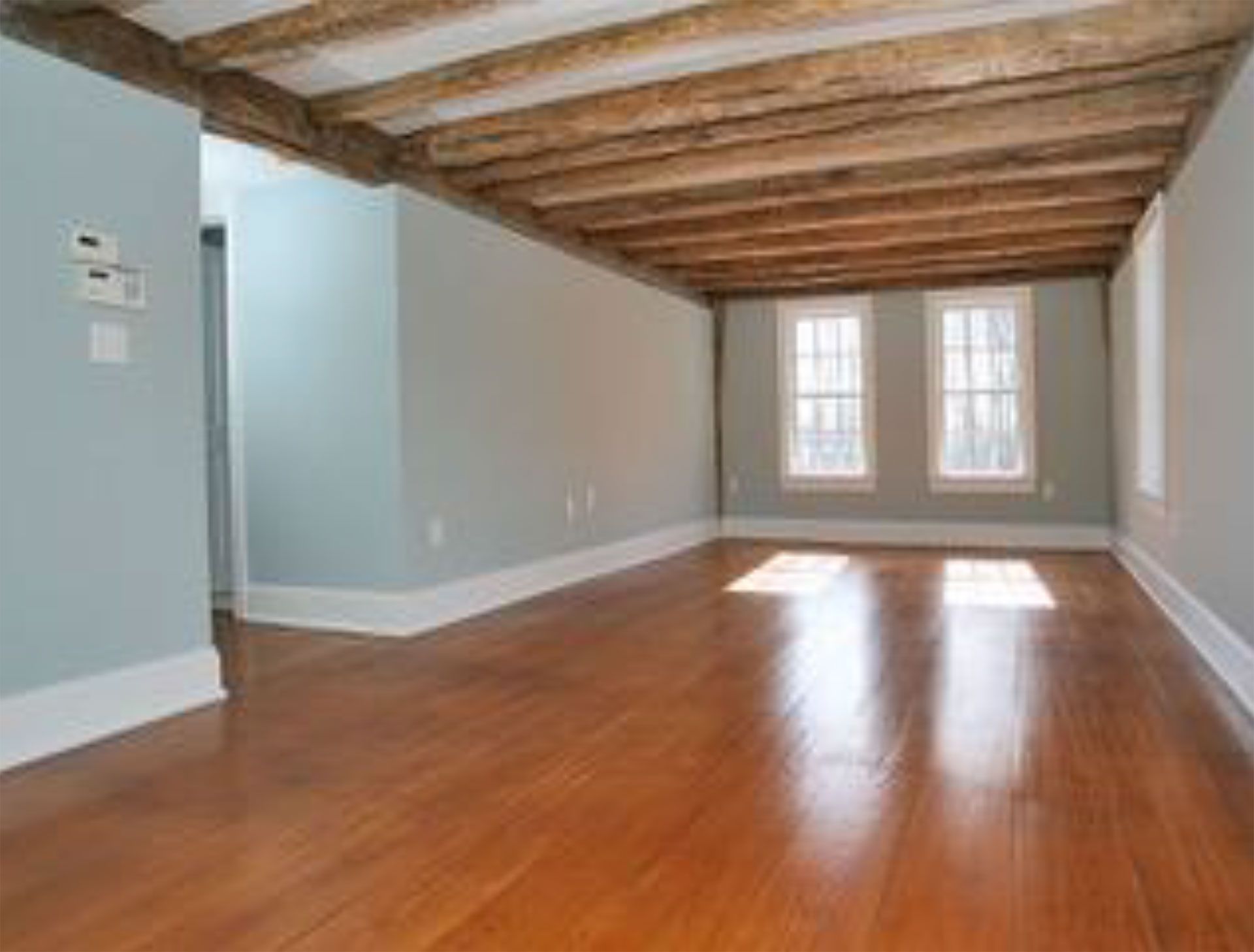 Empty room with hardwood floors, light blue walls, exposed wooden beams, and two windows.