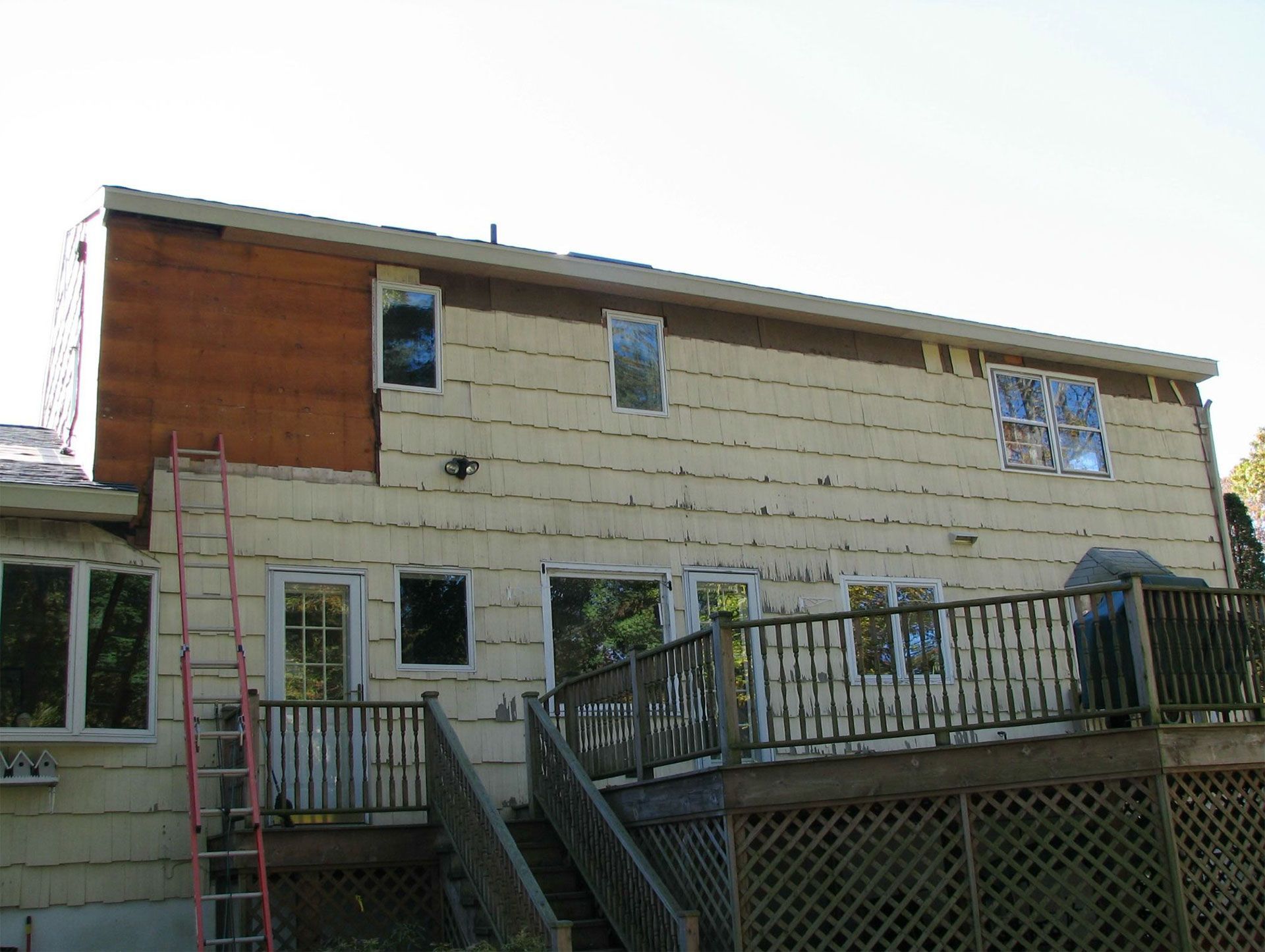 Back of a house being renovated; tan siding, brown section, ladder, deck, windows.