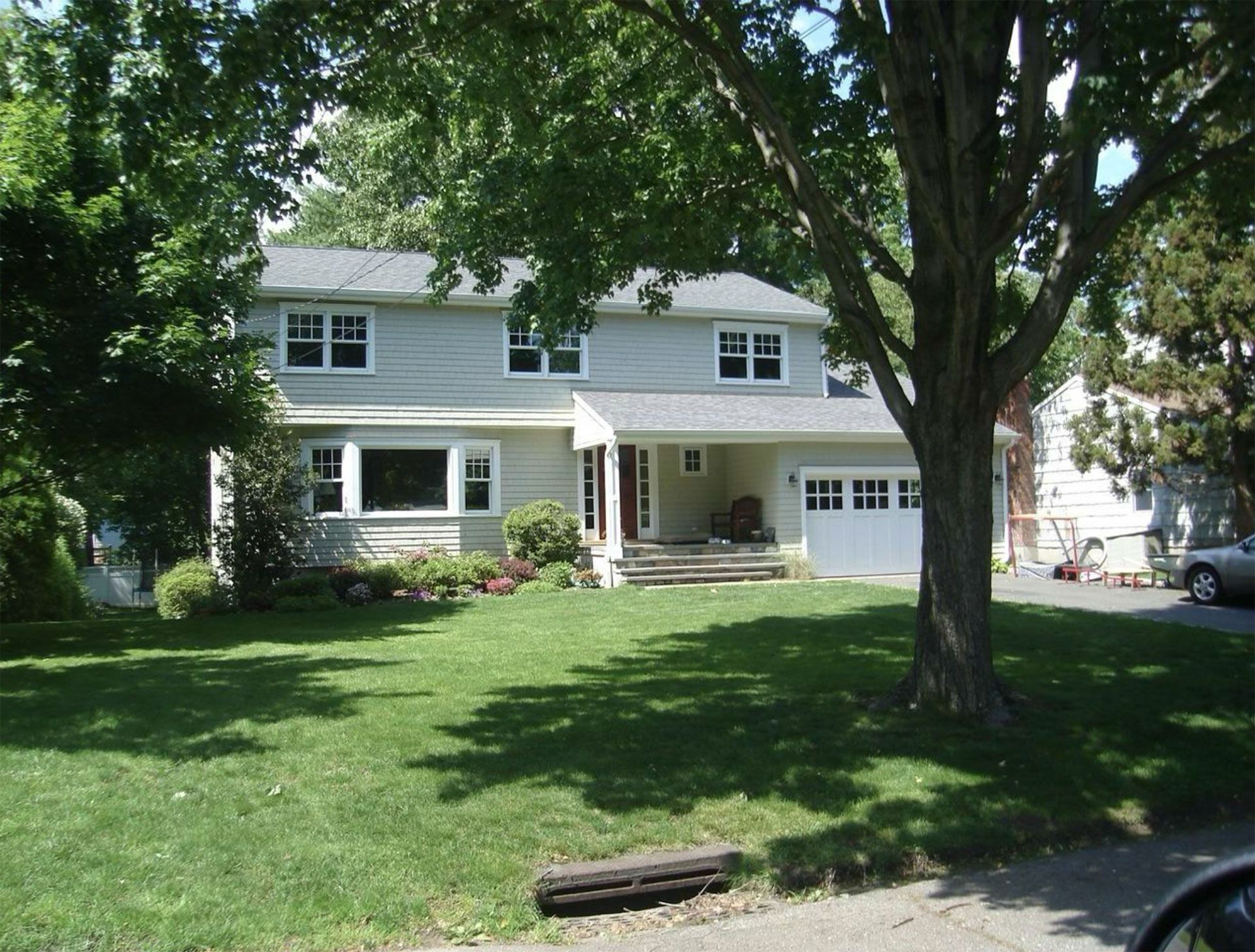 Two-story house with light gray siding, a green lawn, and trees in the front yard. White garage door.