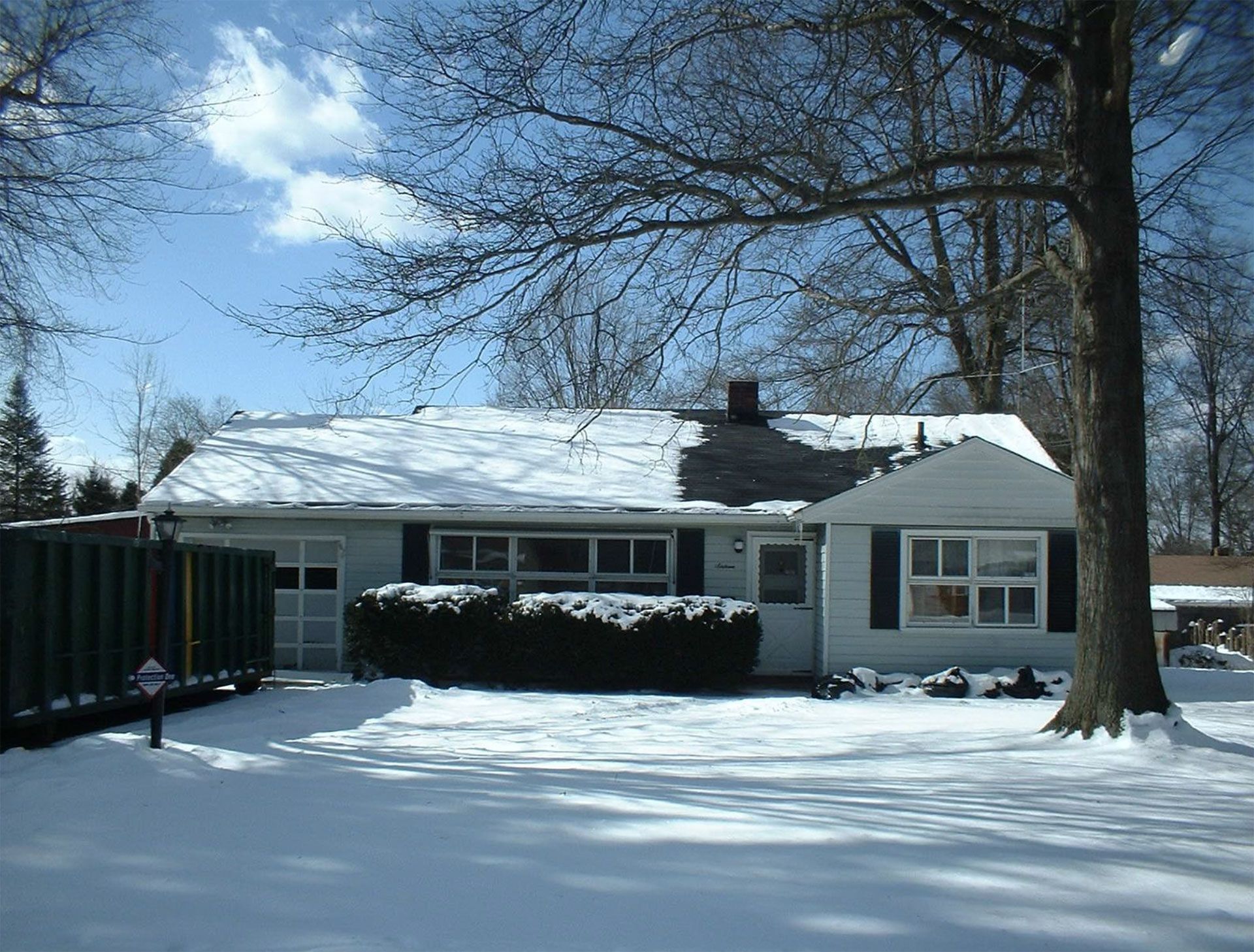 A snow-covered ranch-style home on a sunny winter day. A large tree shades the front yard.
