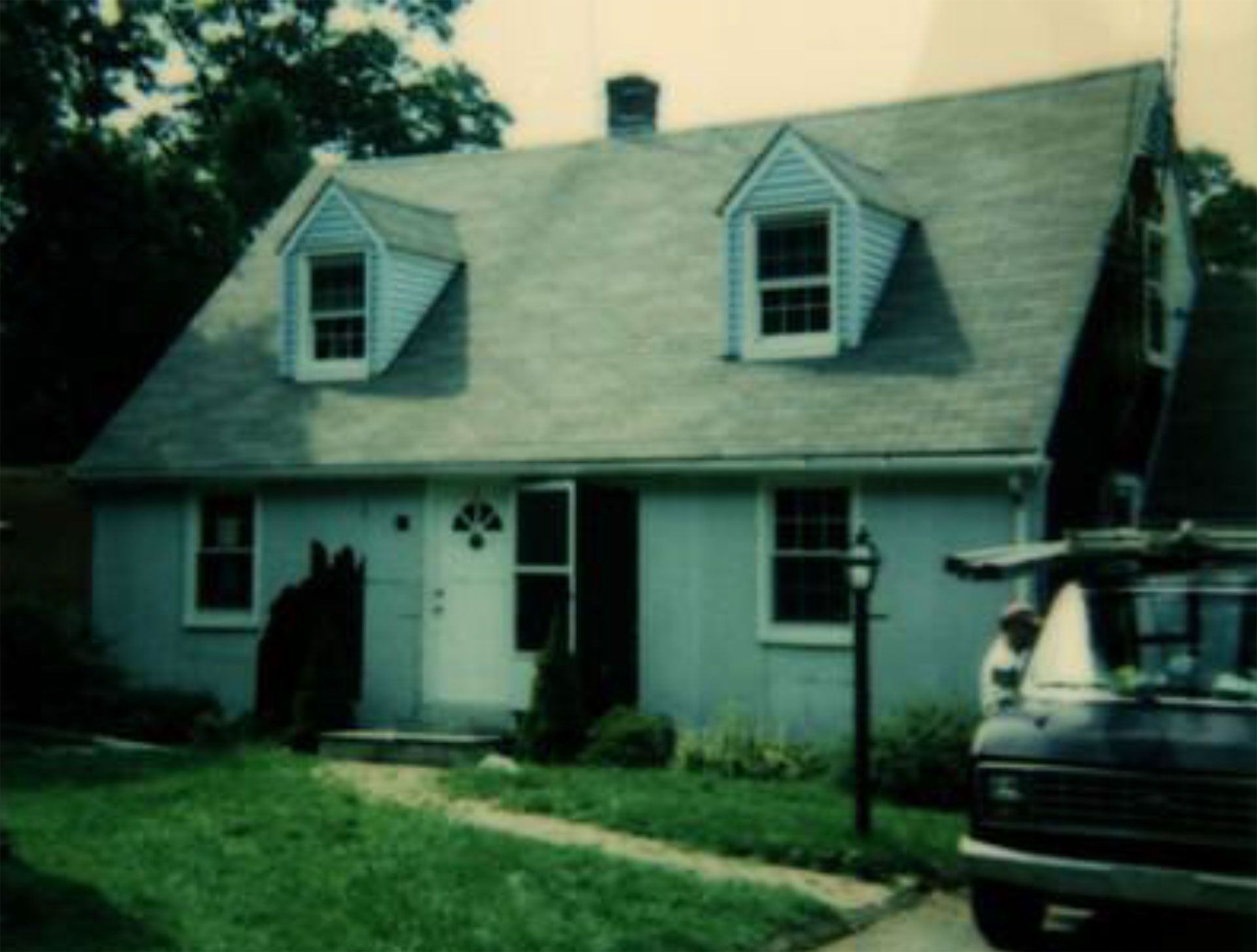 Blue house with dormers, front door, and a vehicle parked in the driveway. Green lawn and trees.
