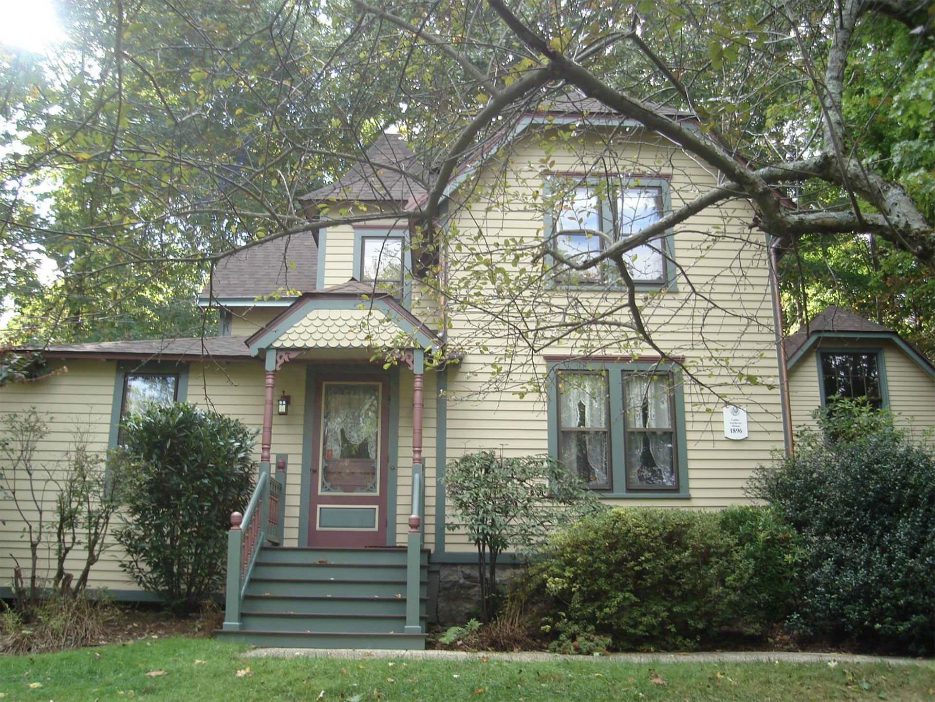 Yellow house with green trim and porch, set amongst trees, sunny day.