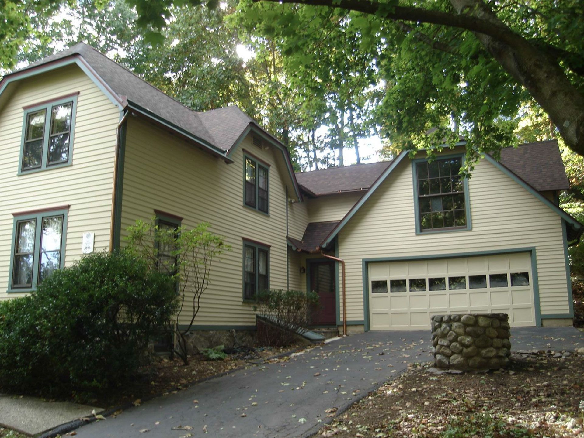 Two-story house with a driveway and attached garage, surrounded by trees. Beige siding, green trim, and brown roof.