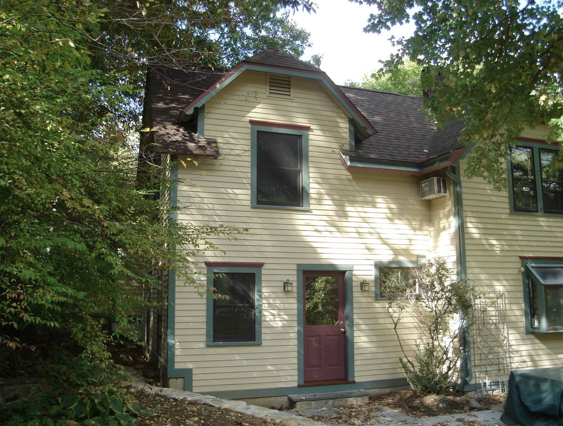 Two-story yellow house with brown roof and burgundy door, surrounded by trees.