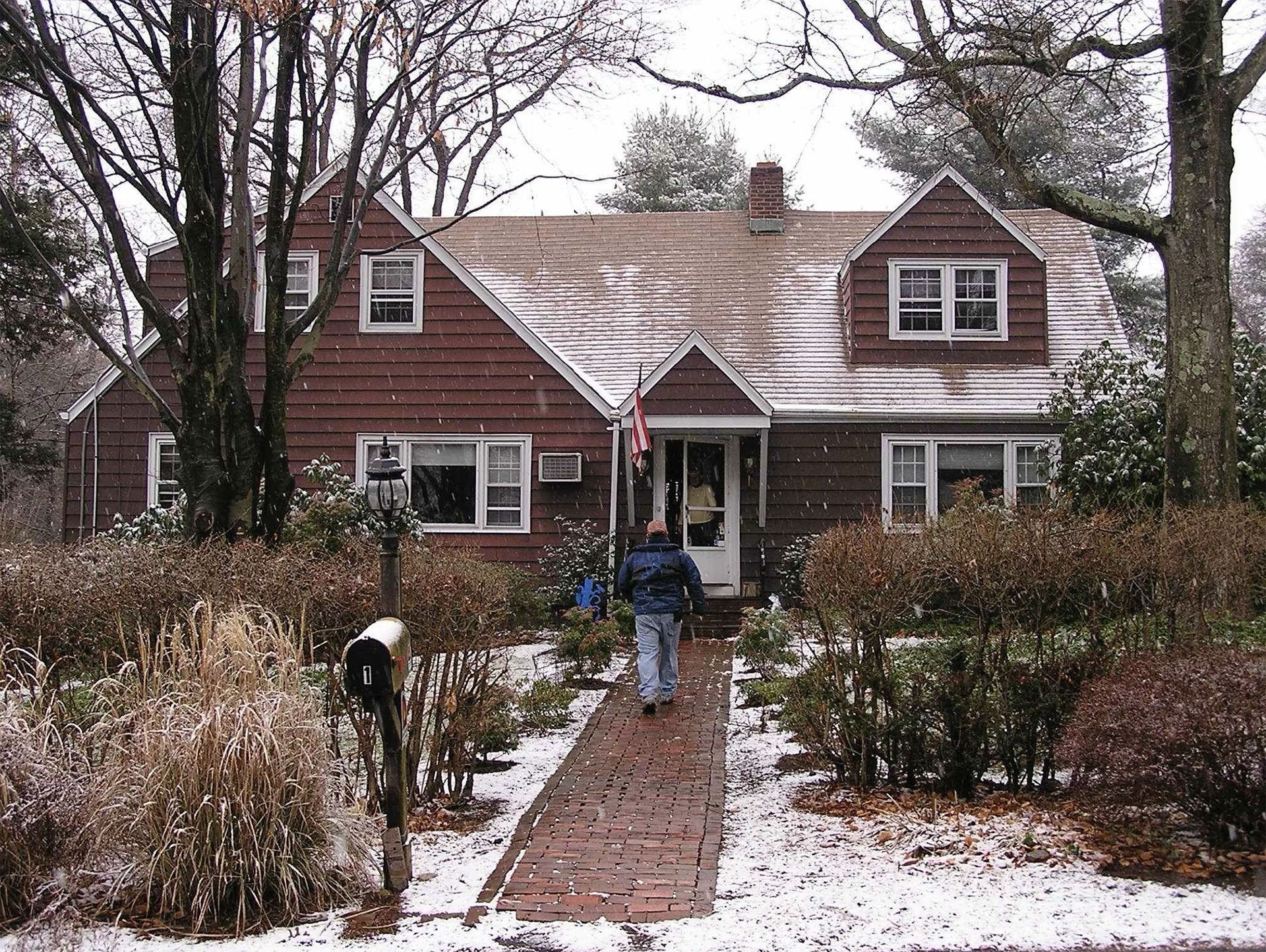A person walks toward a brown house with a brick path in winter, snowy landscape.