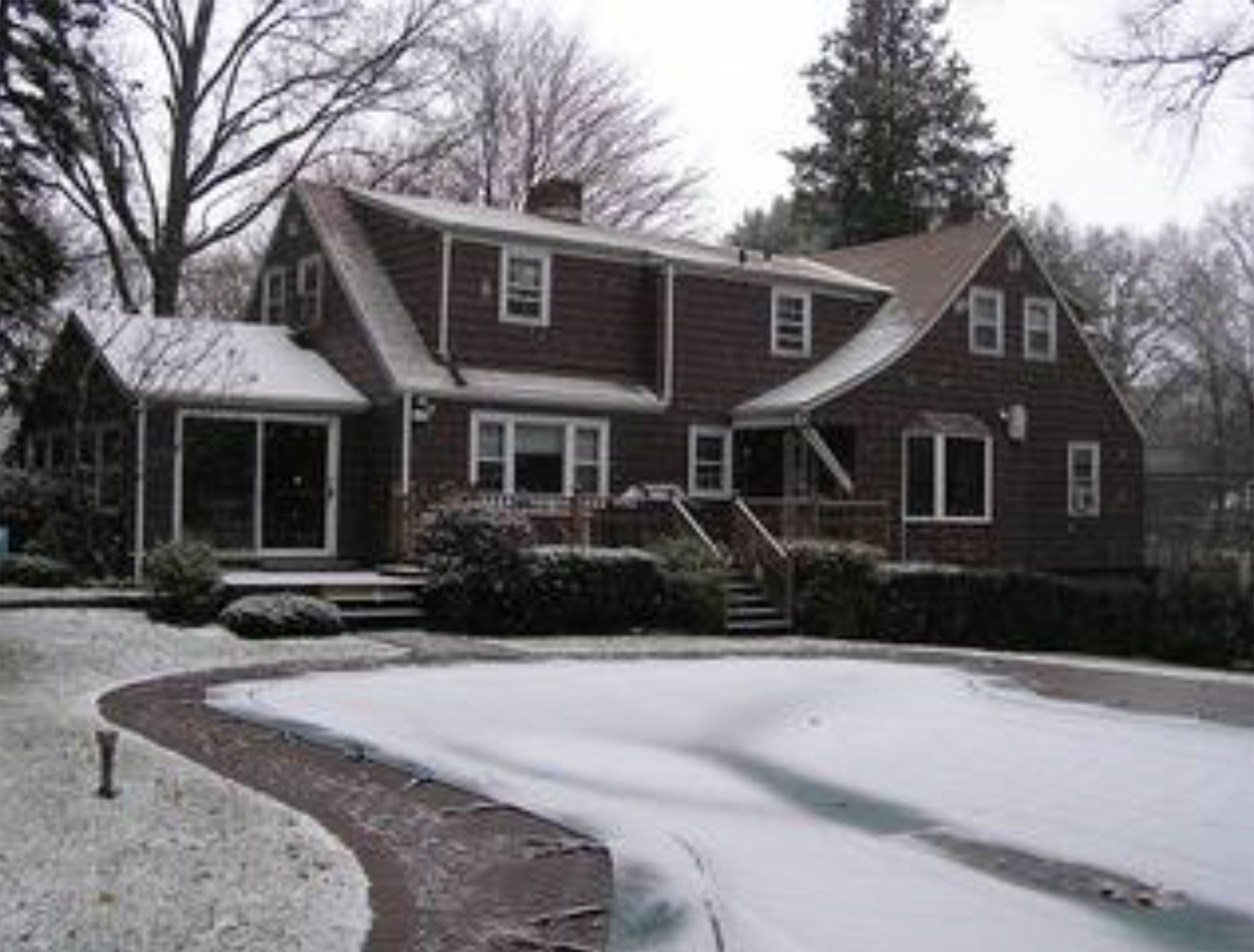 Snow-covered suburban house with dark siding, curved walkway, and bare trees in winter.