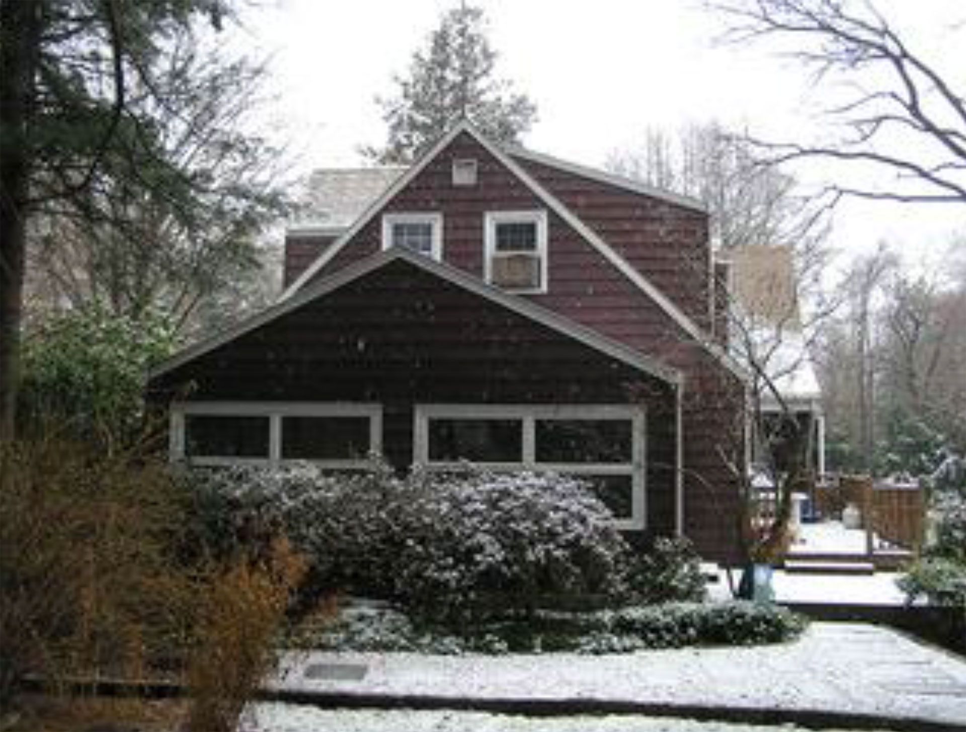 A snow-covered two-story house with dark siding and multiple windows in a wooded setting.