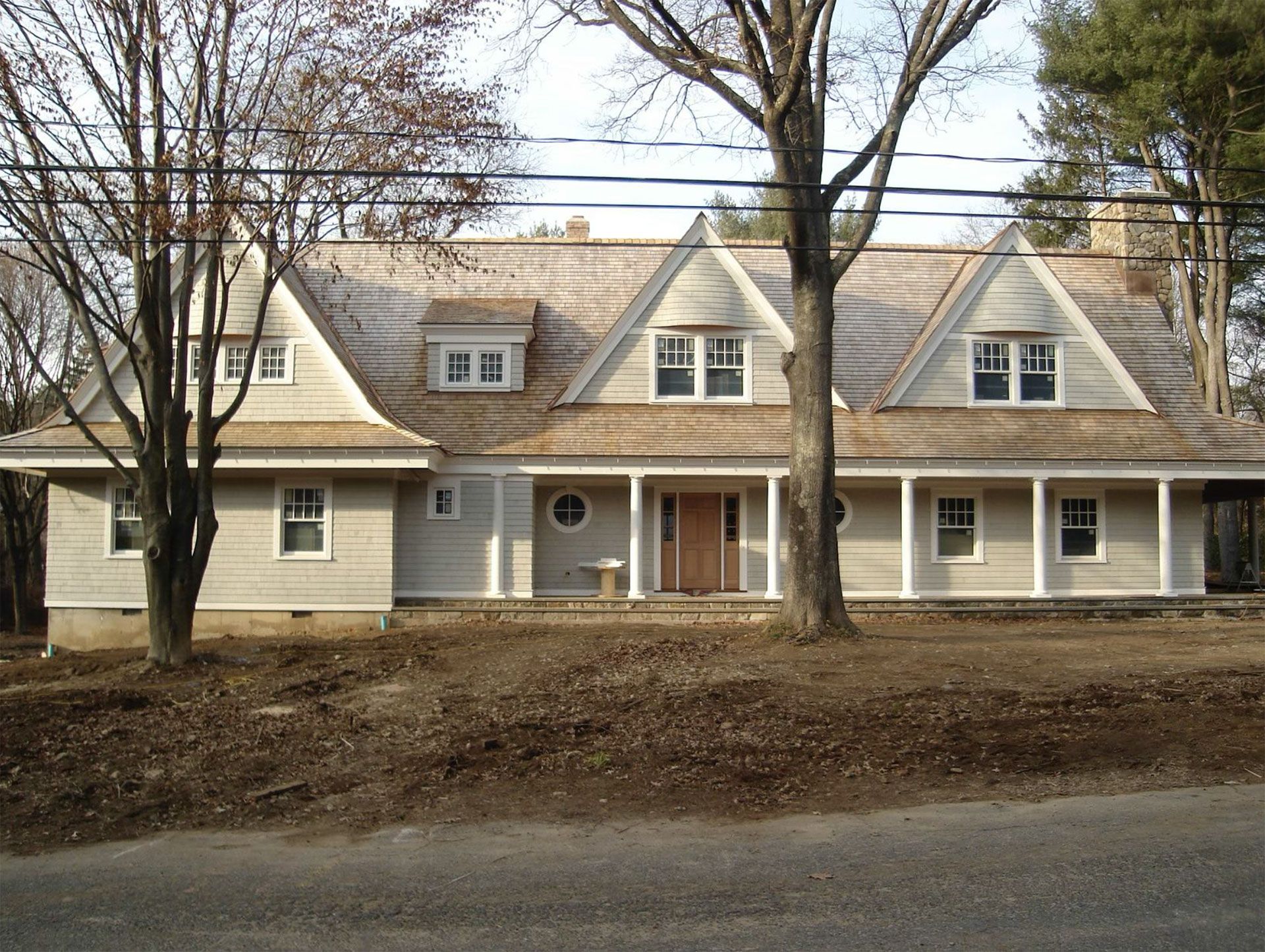 Beige house with porch, gabled roof, and wooden shingles, flanked by trees.