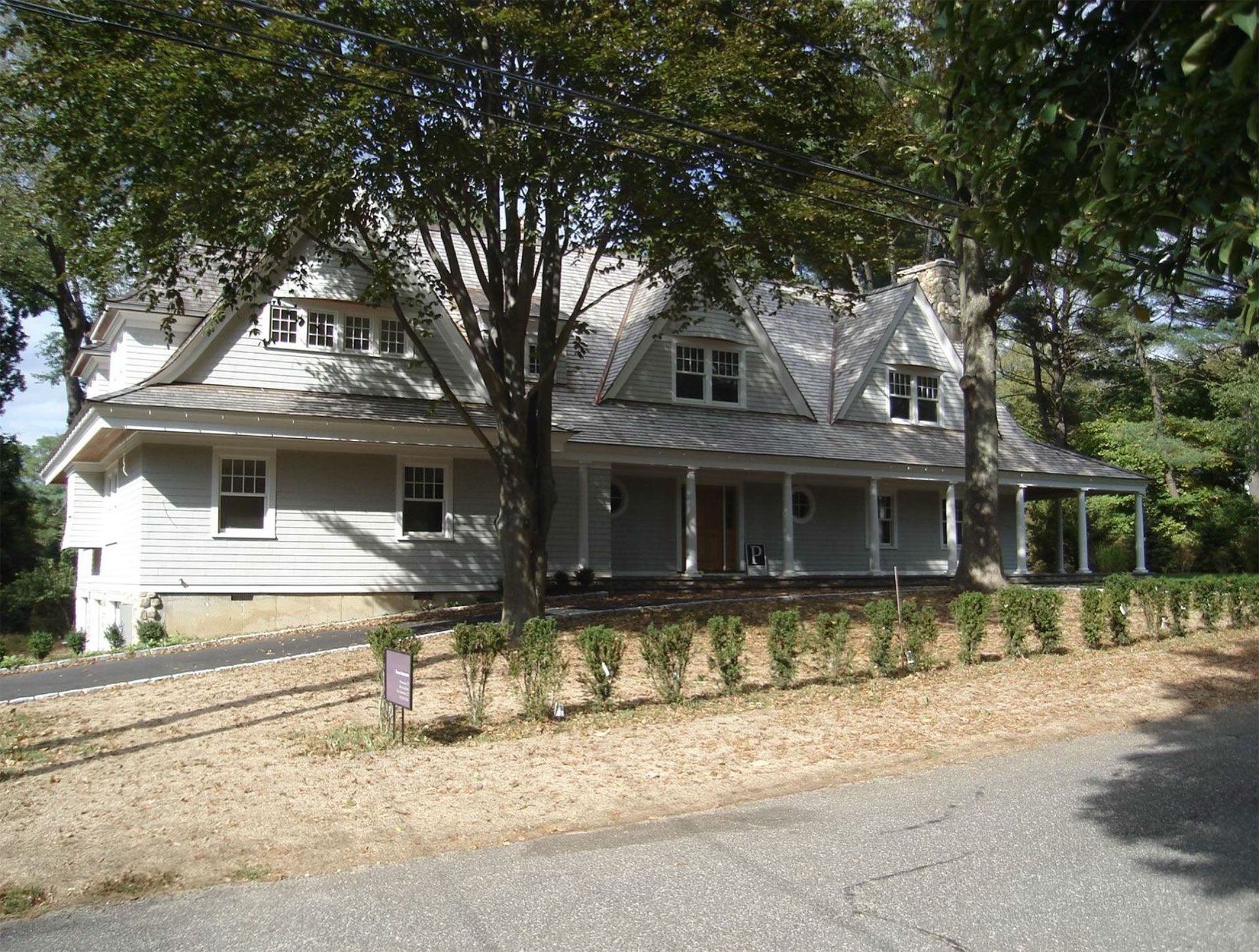 Light-colored house with gray roof, a long porch, and landscaping. Trees and dry ground are in the foreground.