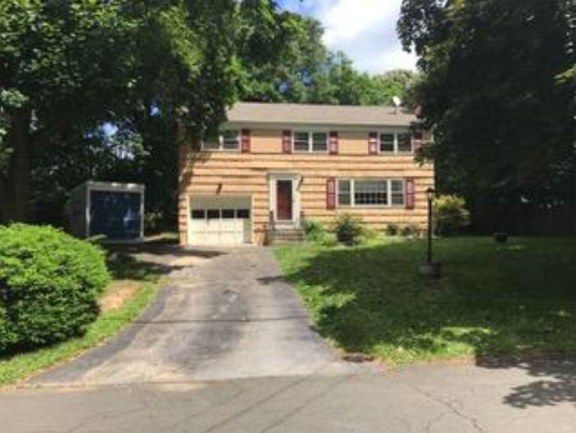 Two-story house with beige siding, red shutters, and a driveway. Trees and a small blue storage container are visible.