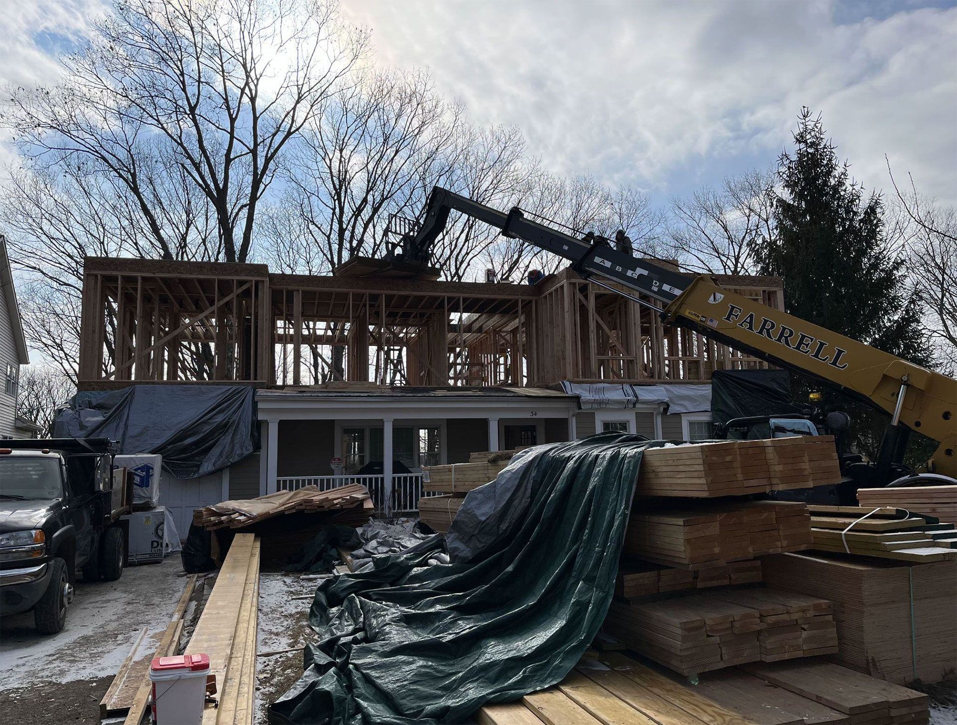 Construction site: framed house addition with crane lifting materials. Lumber pile and truck in front.