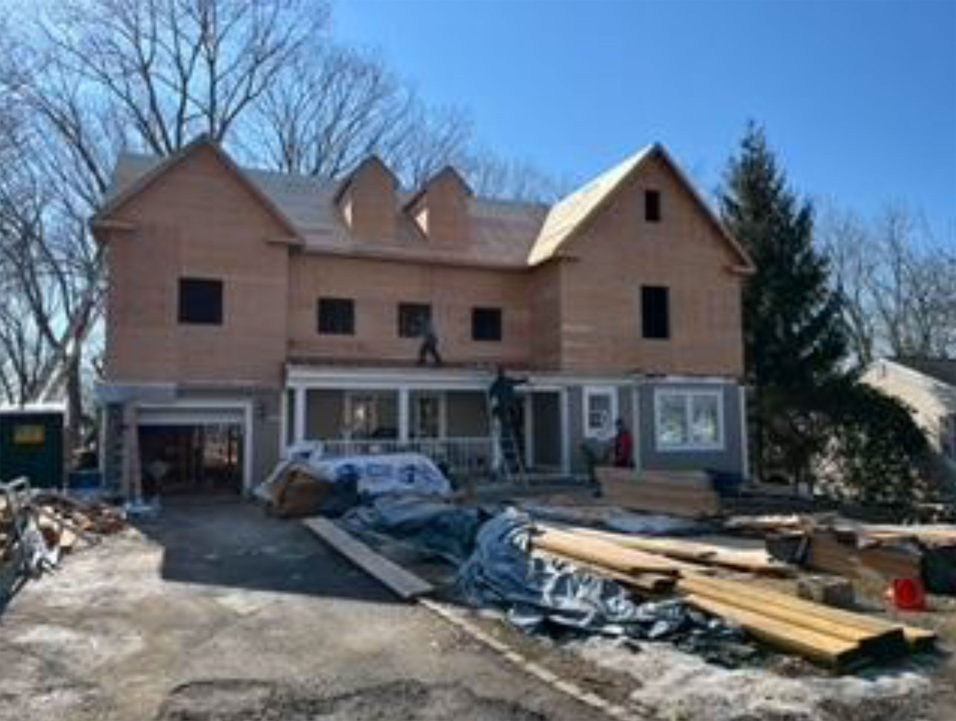 House under construction: wooden frame, workers on roof, building materials on the ground, sunny day.