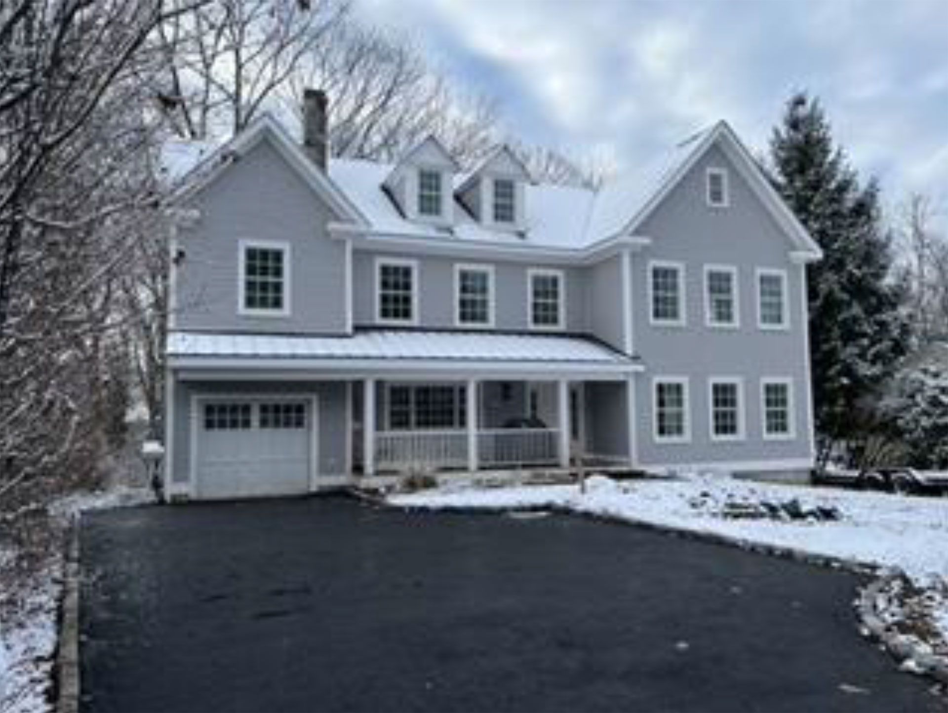 Gray house with snow-covered roof and driveway, featuring a covered porch and attached garage.