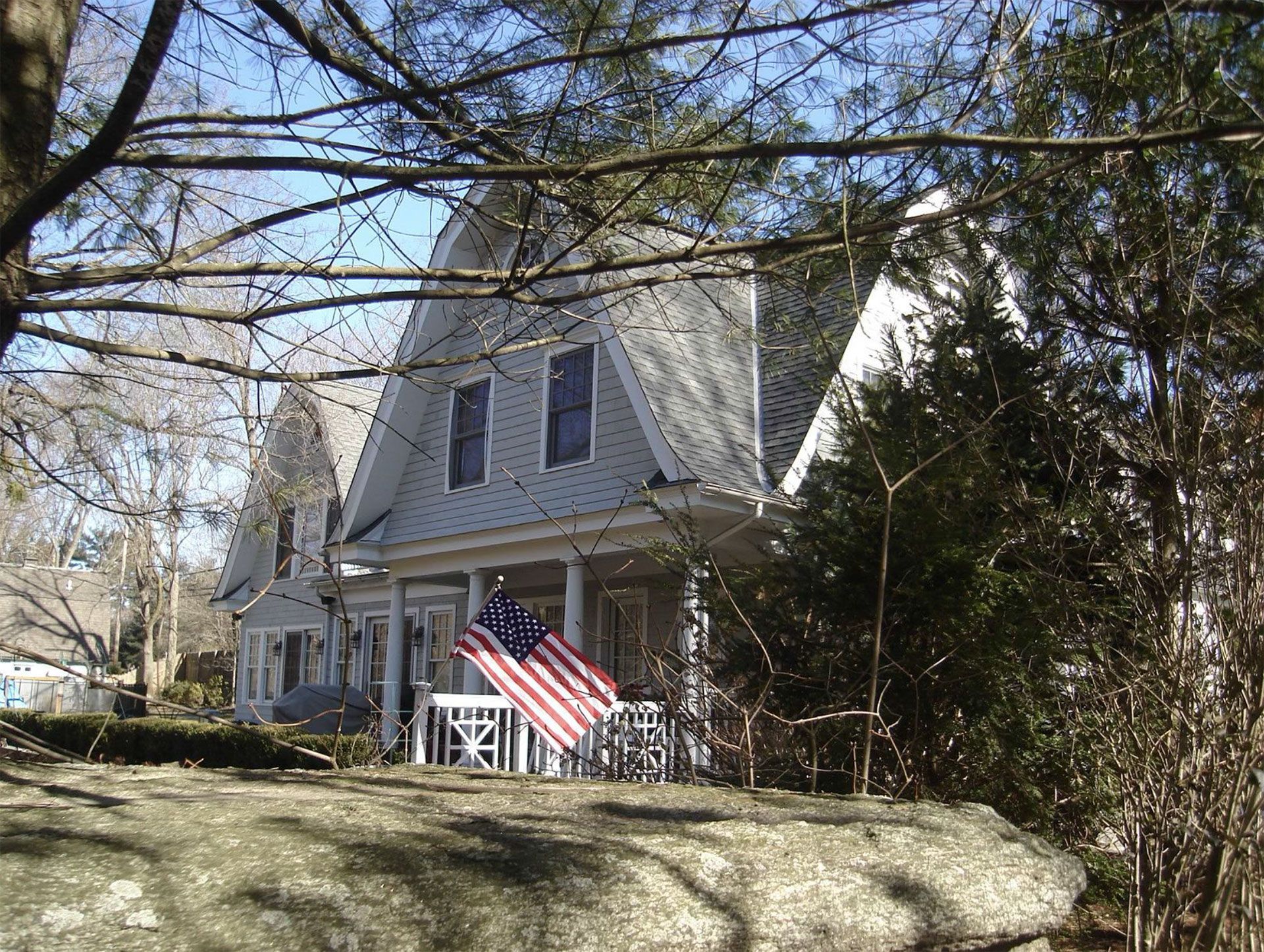 Gray house with a porch, American flag, behind a stone wall, trees in background.