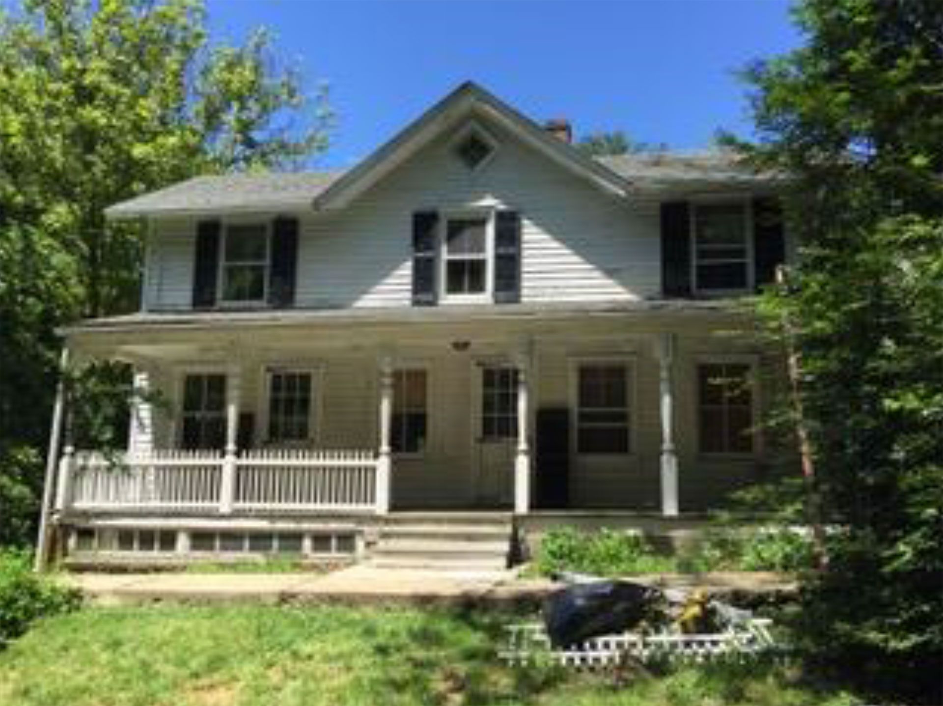 White two-story house with porch and black shutters, trees surround the house, sunny day.